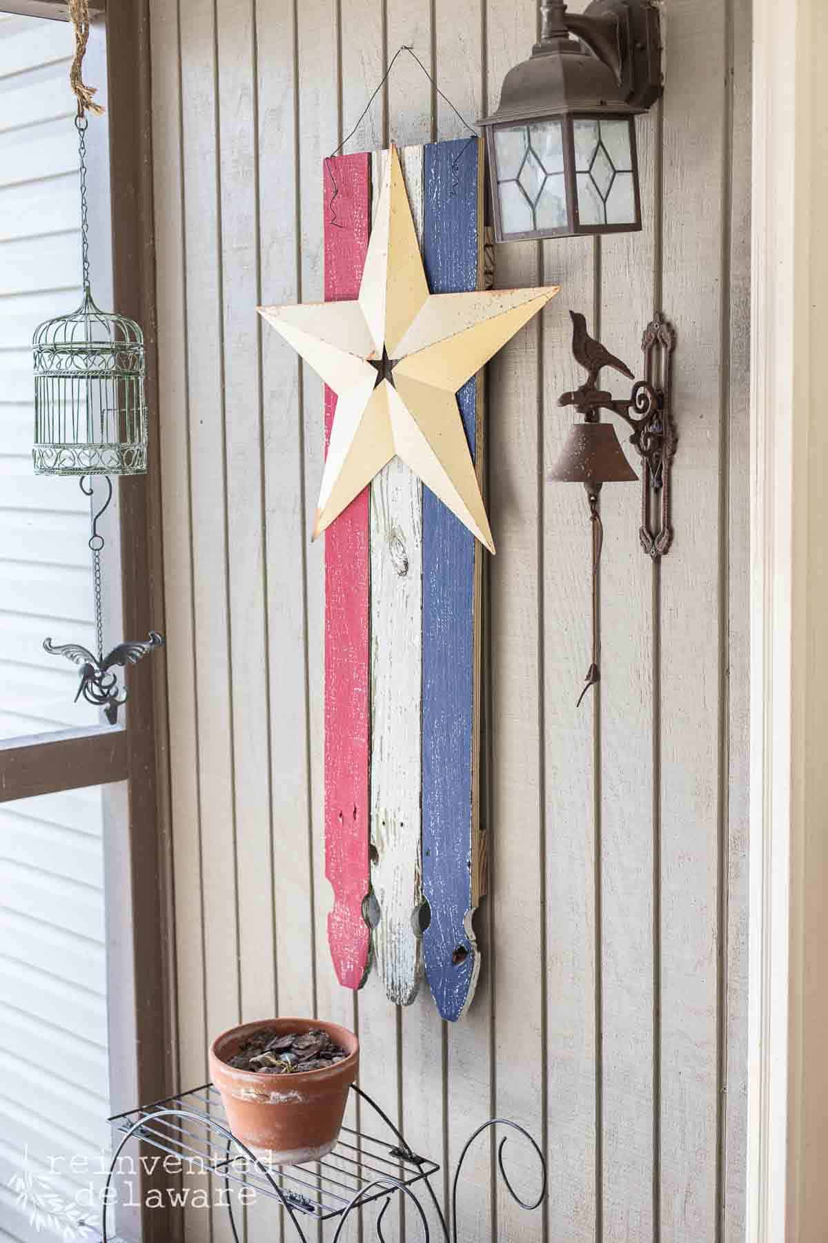 A rustic wood American flag made from reclaimed fence boards with a large yellwo metal star hanging on a back patio for patriotic decor.