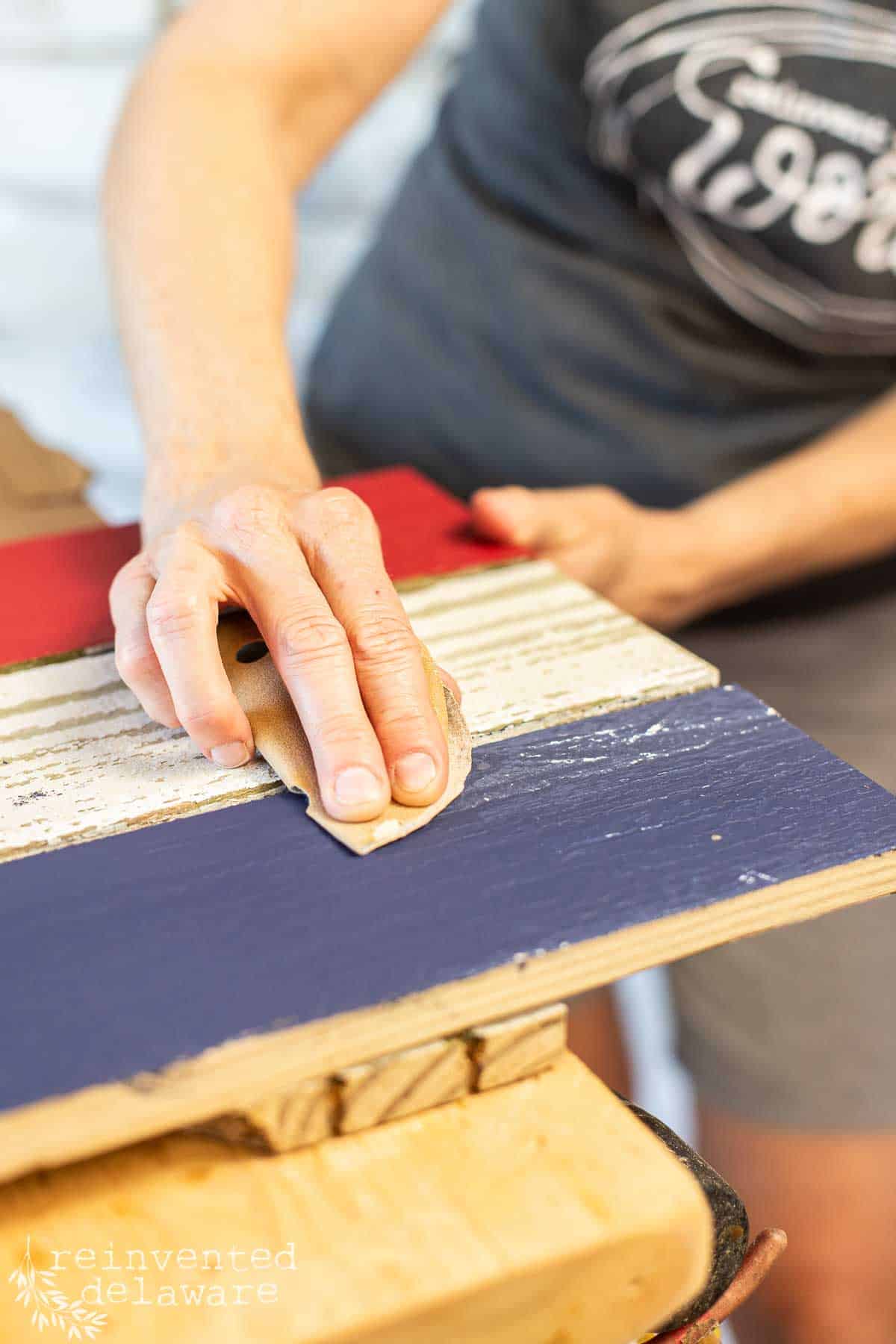 A lady using sandpaper to distress the red, white and blue paint on a handmade wooden American flag.