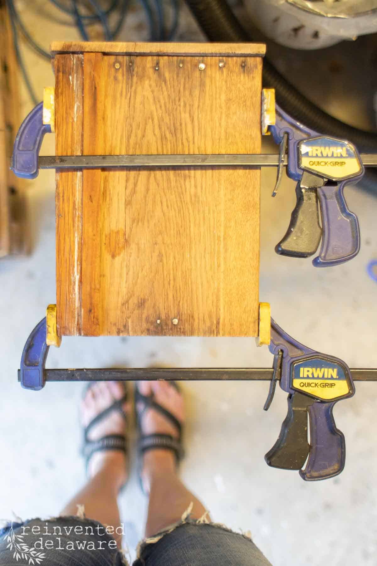 A lady using wood glue and clamps to repair an old drawer on a piece of broken wood furniture.