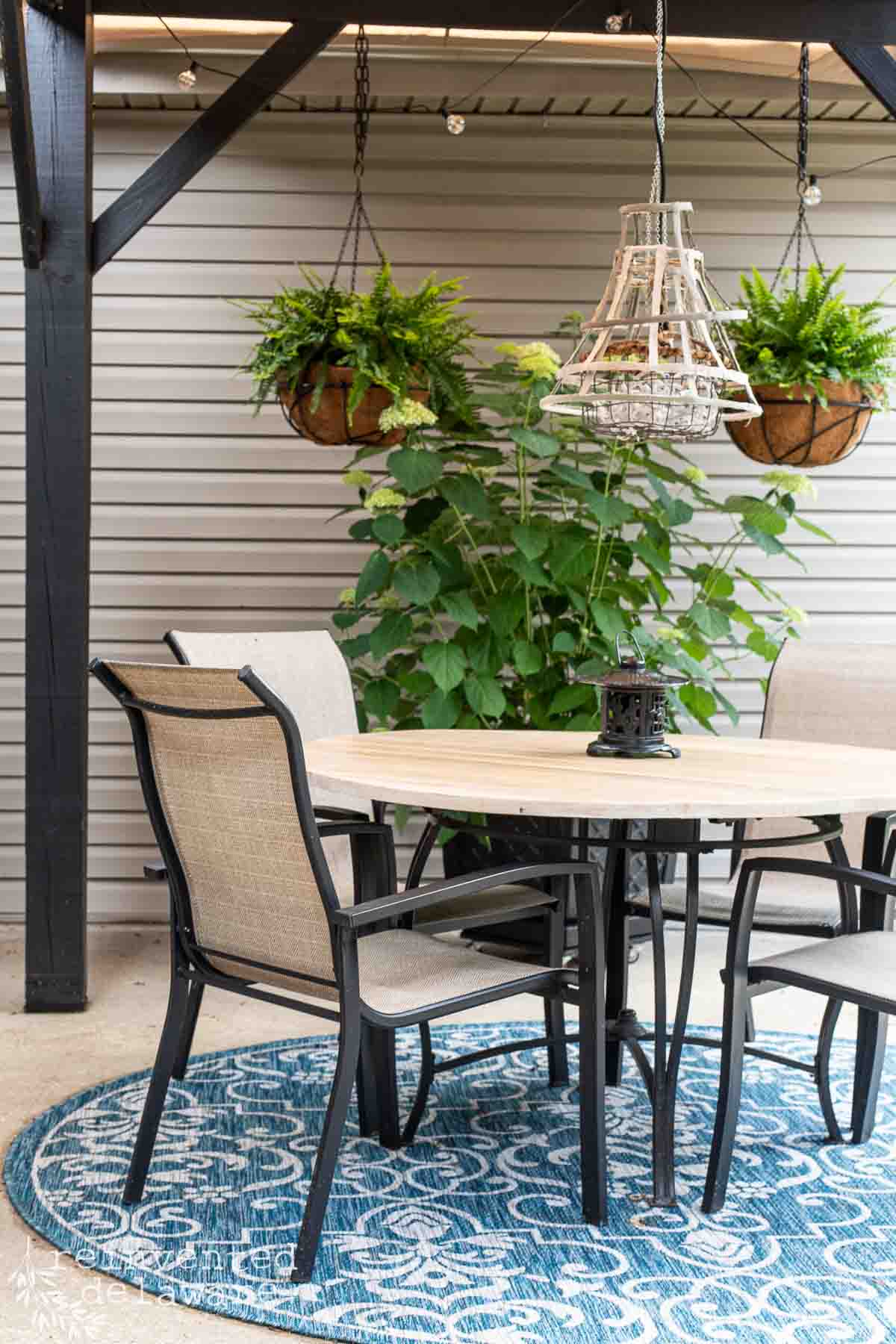 Shady area on a cement patio with a pergola that has a diy sun shade attached to it. Hanging baskets with ferns and a potted hydrangea for outdoor decor.