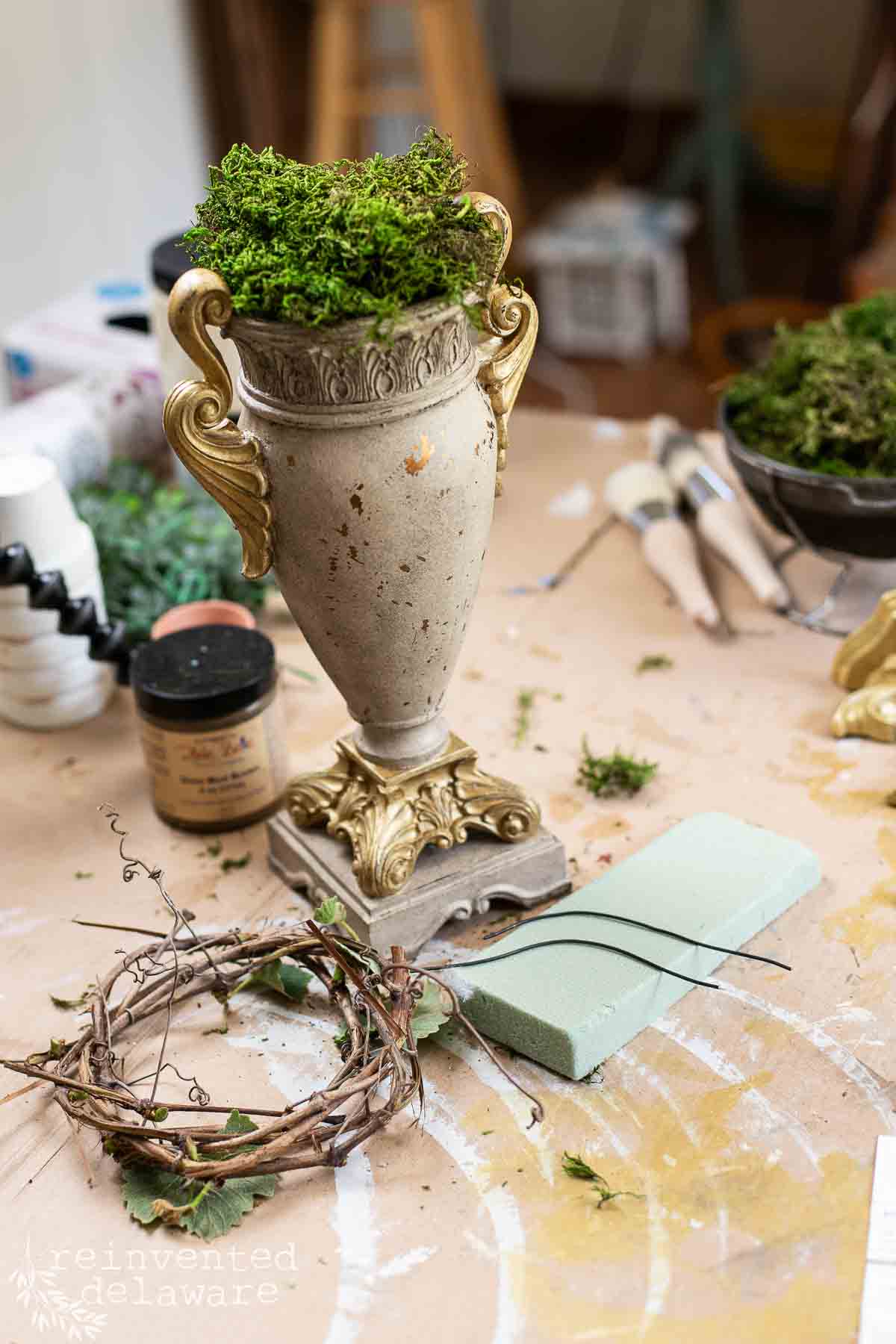 Supplies used to upcycle an old urn from the thrift store incluidng paintbrushes, paint, florist foam and a handmade grapevine wreath.