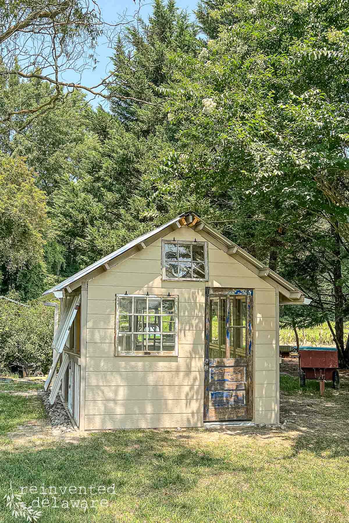 A small, rustic, light-yellow shed with large windows stands surrounded by trees on a sunny day. The shed has a wooden door and a glass design near the roof peak. A wheelbarrow is visible to the right of the shed.