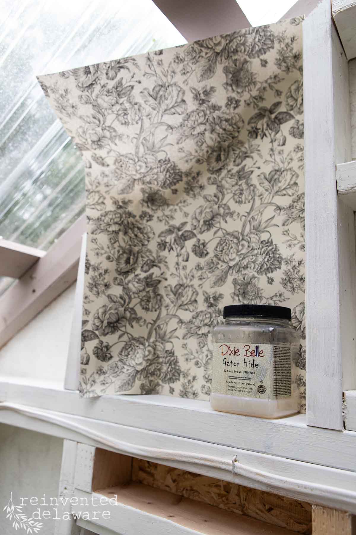 A jar of Dixie Belle Gator Hide sealer sits on a white wooden ledge in front of a partially installed sheet of floral-patterned drawer liner paper inside a greenhouse with glass windows. The wallpaper has a black-and-white flower design. .
