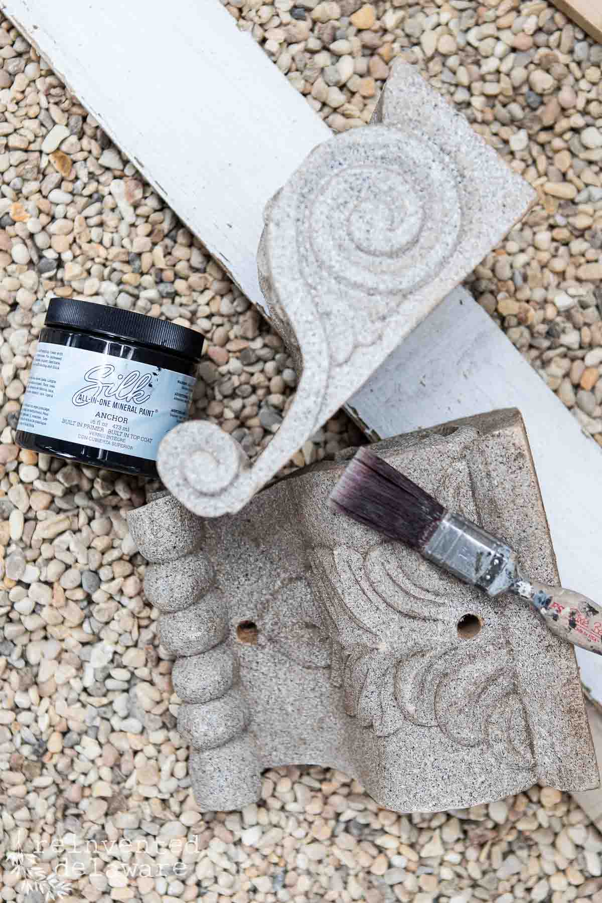 A jar of Dixie Belle Silk chalk paint, a paintbrush with residue, and two ornate, unfinished corbels lie on top of a gravel surface. The corbels have intricate carvings and are partially painted.