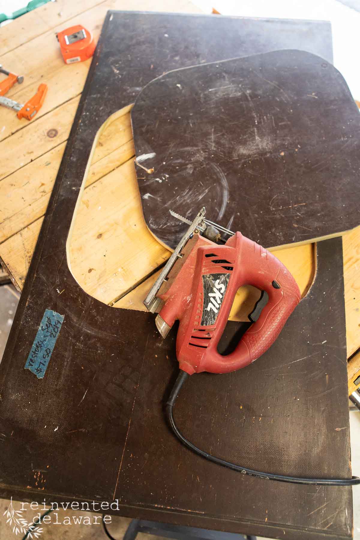 A red jigsaw tool rests on a cut-out wooden board, partially placed on a light wooden table. In the background, a tape measure, ruler, and orange clamp are visible. The edges of the wooden board are round and smoothed, indicating ongoing woodworking activity.