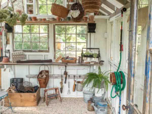 A quaint potting shed with large windows letting in natural light. The workspace is lined with gardening tools, pots, and various supplies. Hanging baskets and potted plants adorn the space, showcasing charming garden shed ideas for a rustic interior.