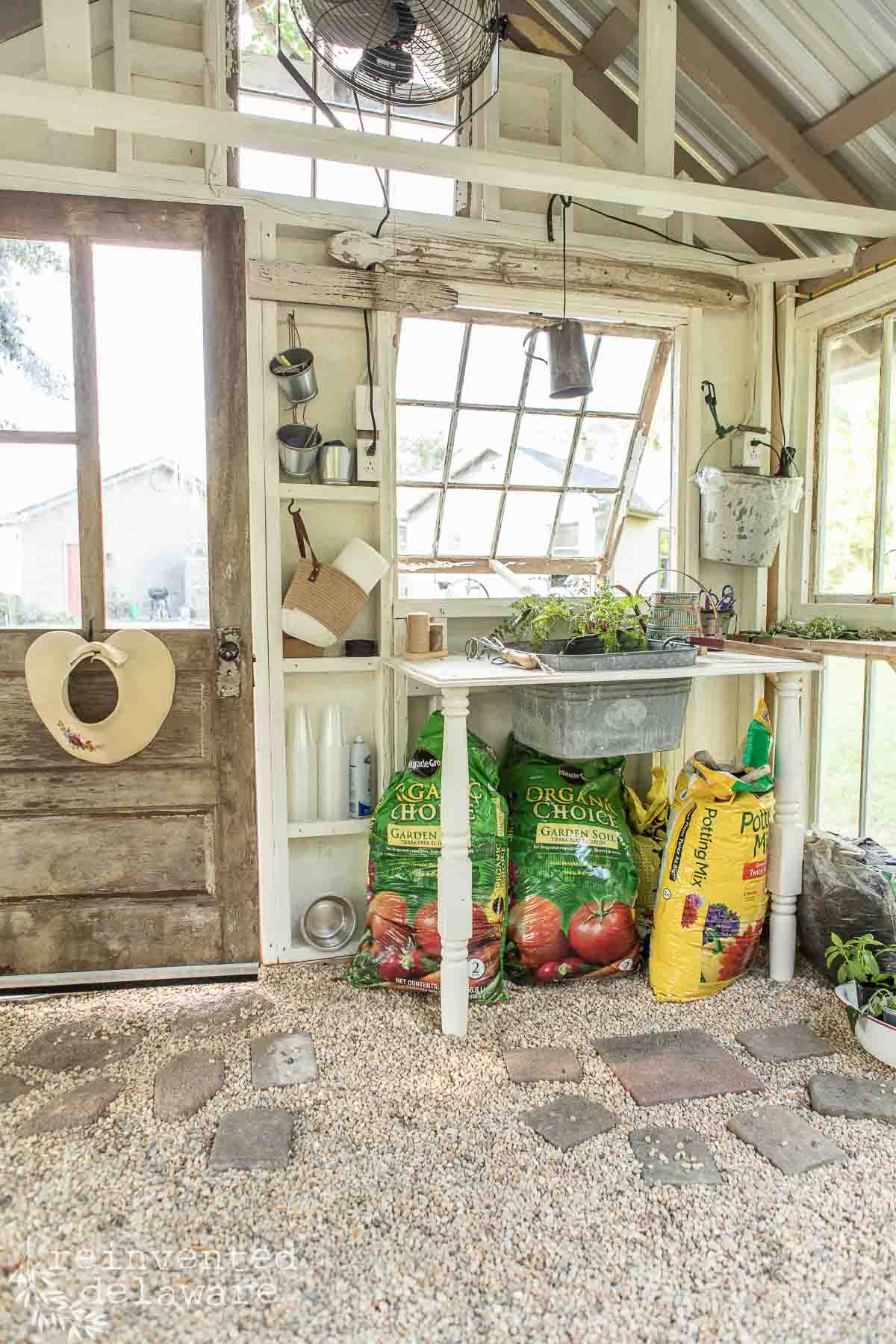 A quaint greenhouse interior with a rustic wooden door on the left. Inside are two large bags of soil, gardening tools, a table with potted plants, and a faucet with a basin. Seedlings are on the counter under a tilted window, and the floor is covered with small gravel.