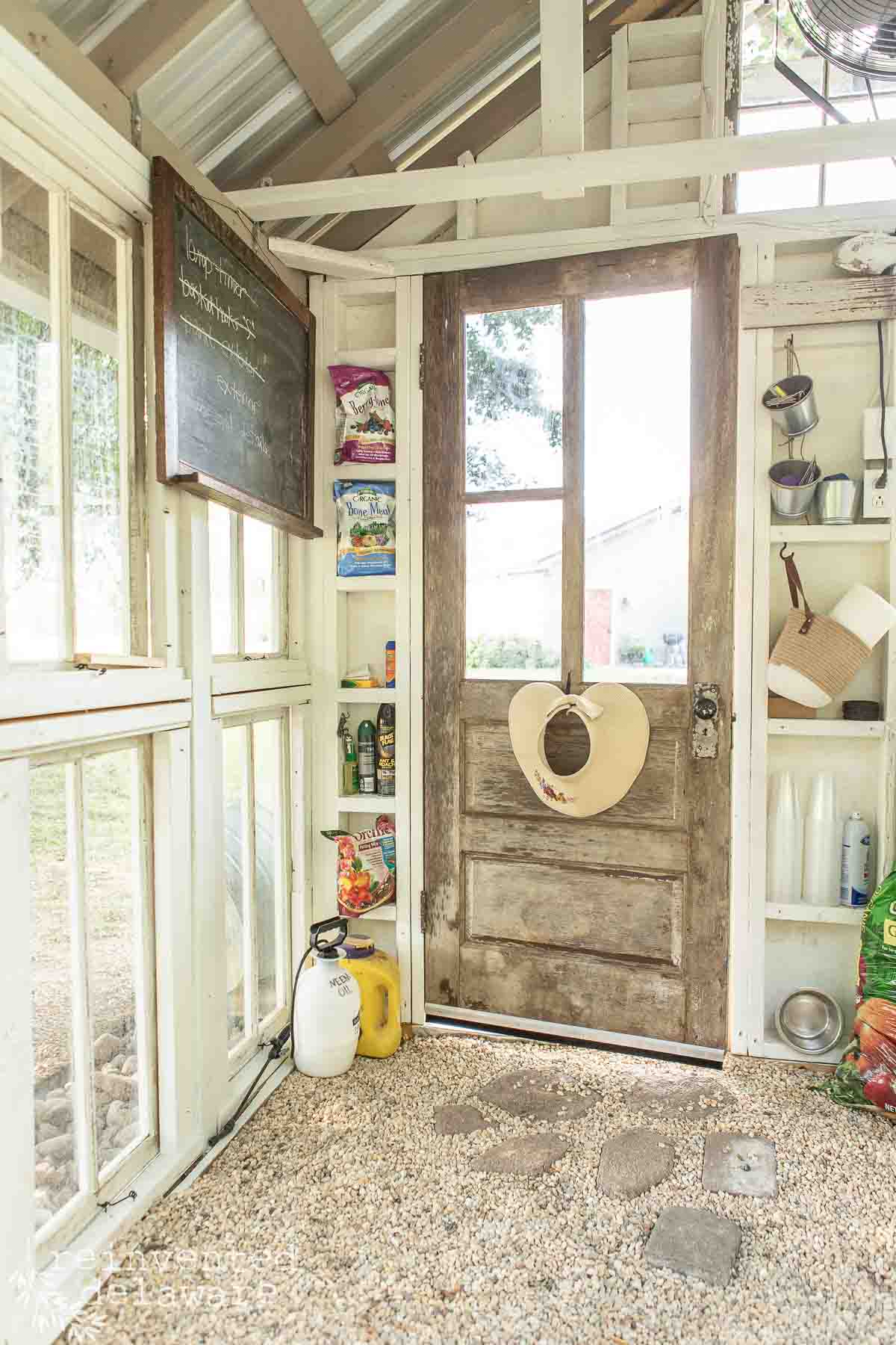 Rustic wooden door in a bright, greenhouse-like room with shelves holding gardening supplies, bug spray, and a chalkboard on the left with a list. A beige hat hangs on the door. Gravel floor and a view of greenery through the windows indicate an outdoor setting.