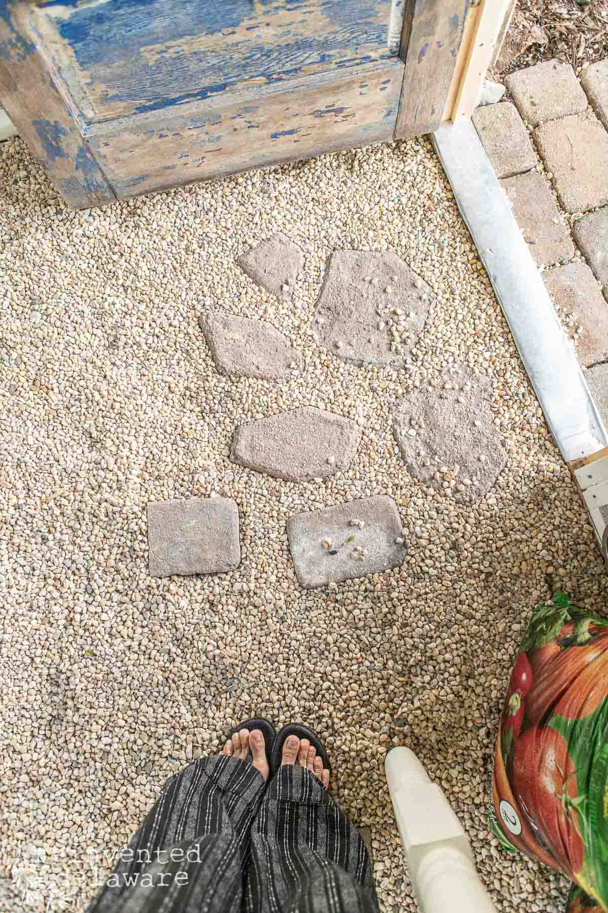 View of garden shed interior with gravel flooring and stepping stones leading to a rustic blue door. Person wearing black flip-flops and striped pants stands in the foreground. To the right, a bag of garden soil is partially visible.