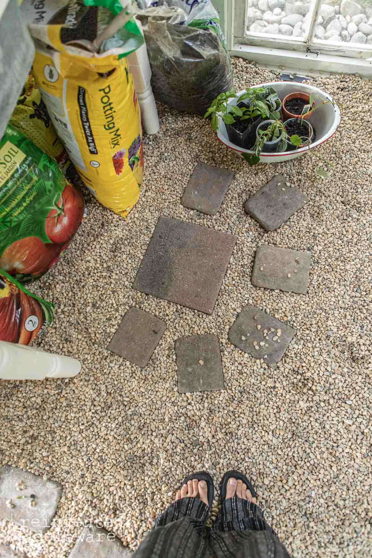 View of a small indoor garden shed with bags of potting soil, a few potted plants, and paving stones arranged on a gravel floor. Feet in sandals and pants are visible at the bottom of the image.