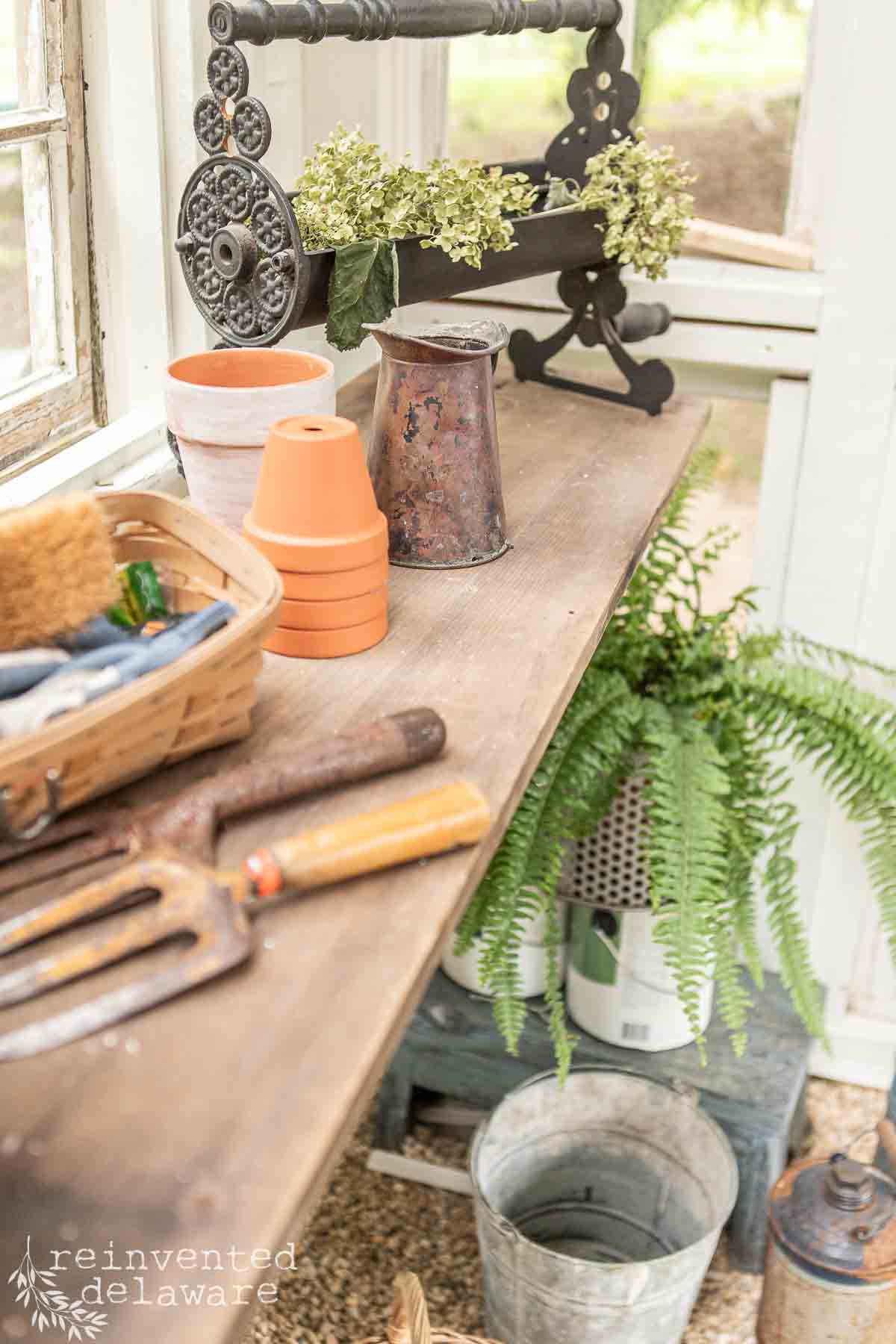 A rustic gardening workstation with a wooden shelf holding garden tools, clay pots, a rusty watering can, and a wicker basket. A metal plant stand with greenery is on the left, and a potted fern sits on the floor below.