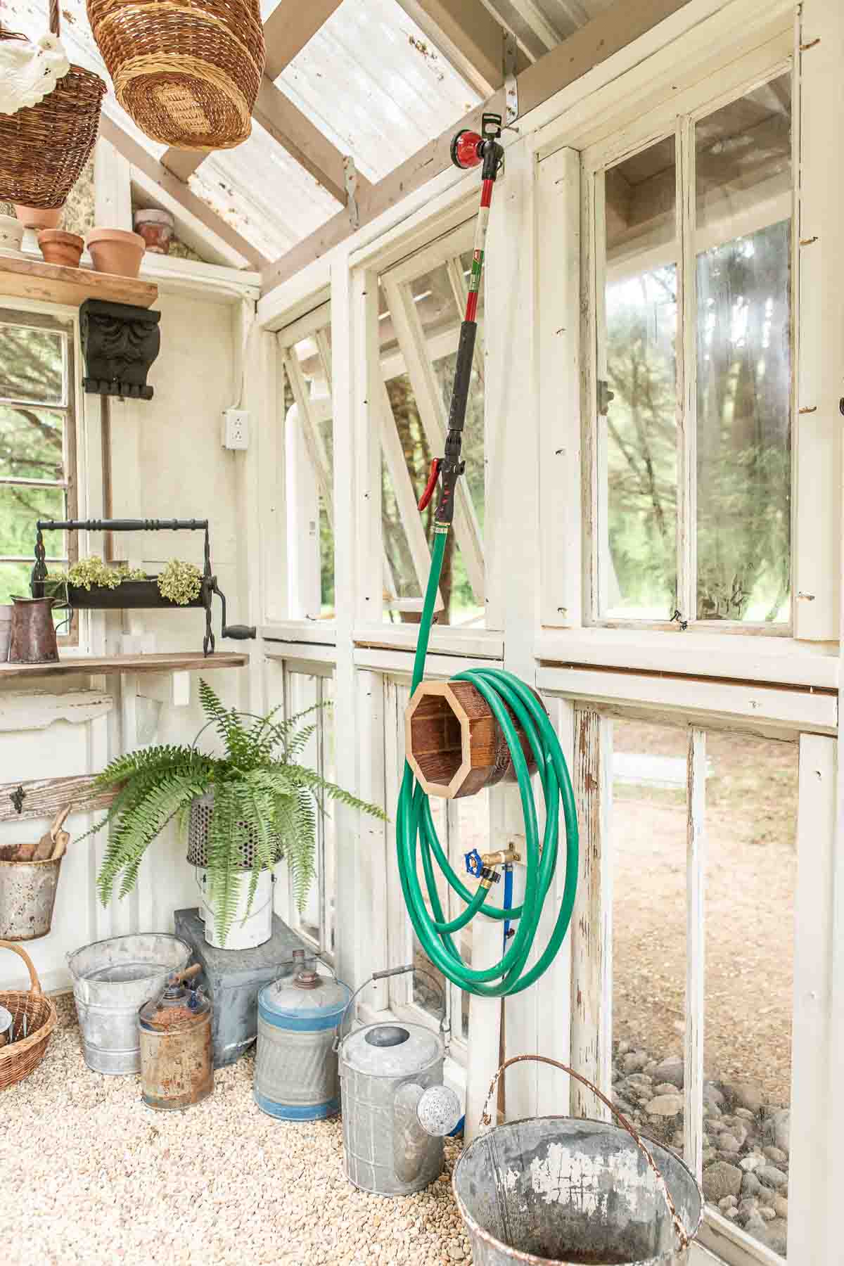 A well-lit greenhouse interior with a coiled green garden hose mounted on the wall beside a telescopic tool. Various gardening tools, potted plants, and metal watering cans are neatly arranged on shelves and the floor. Windows provide a view of greenery outside.