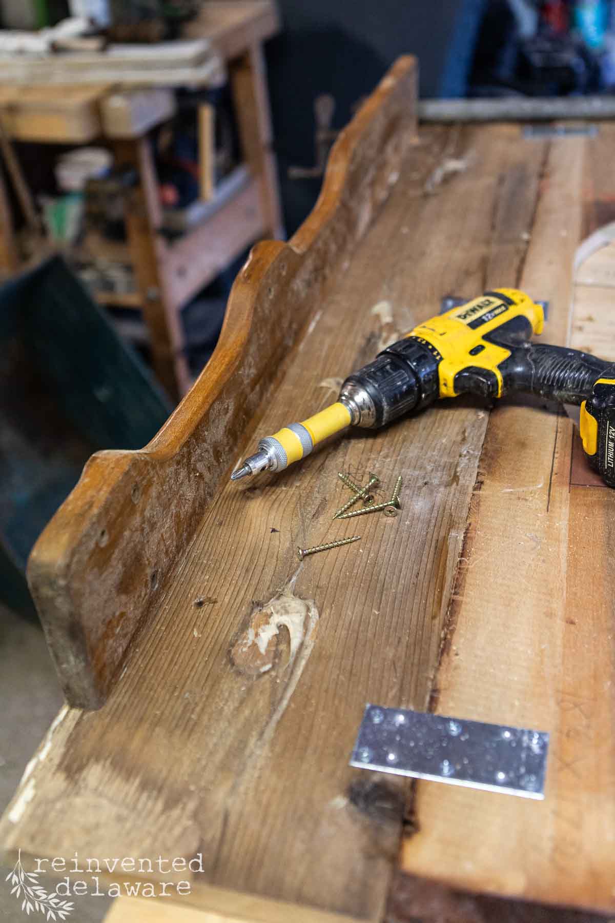 A workbench in the potting shed features a partially assembled wooden shelf, a yellow and black power drill, screws, and a metal bracket. Tools and materials are scattered in the background, suggesting an ongoing DIY project in this cozy workshop.