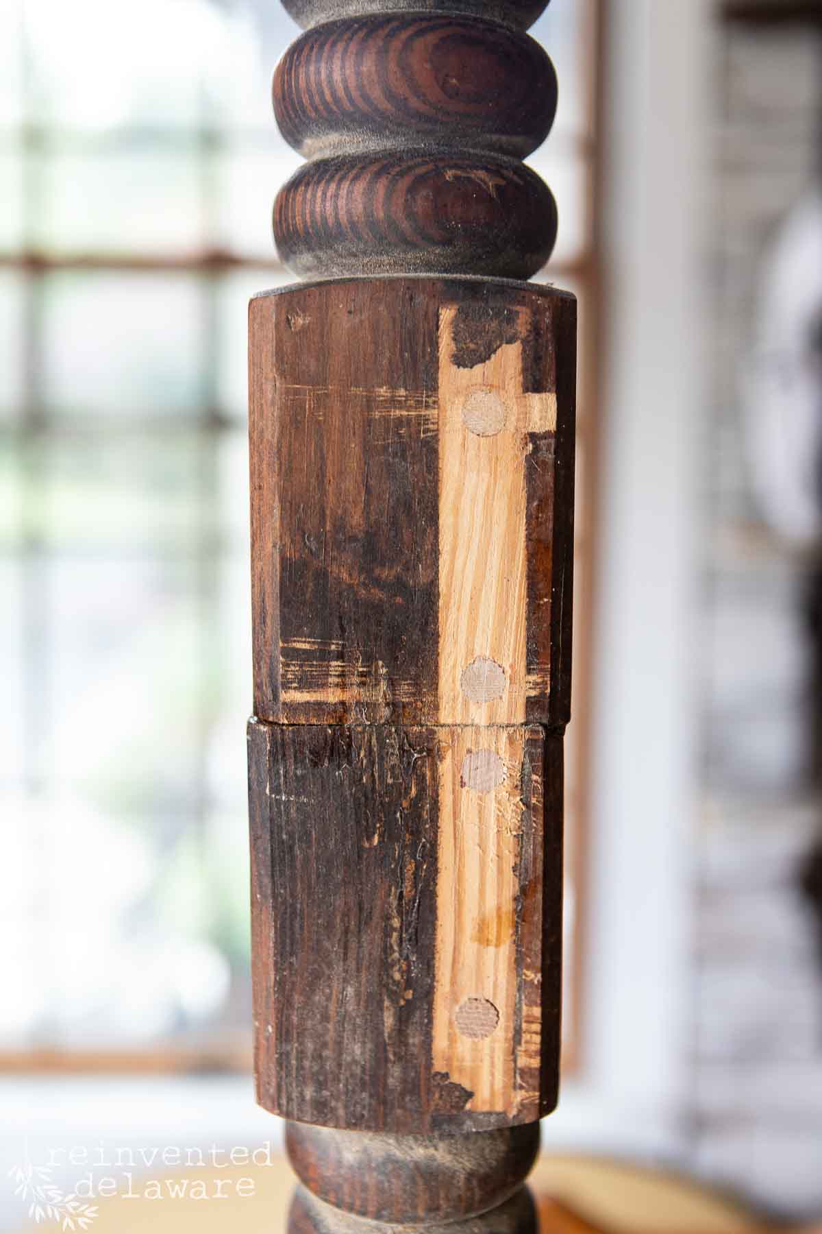 Close-up of a wooden table leg with visible wear and repair. The wood displays a mix of dark and light tones, with a patched section on one side. The leg features turned detailing at the top and bottom. The blurred background shows a window with light coming through.