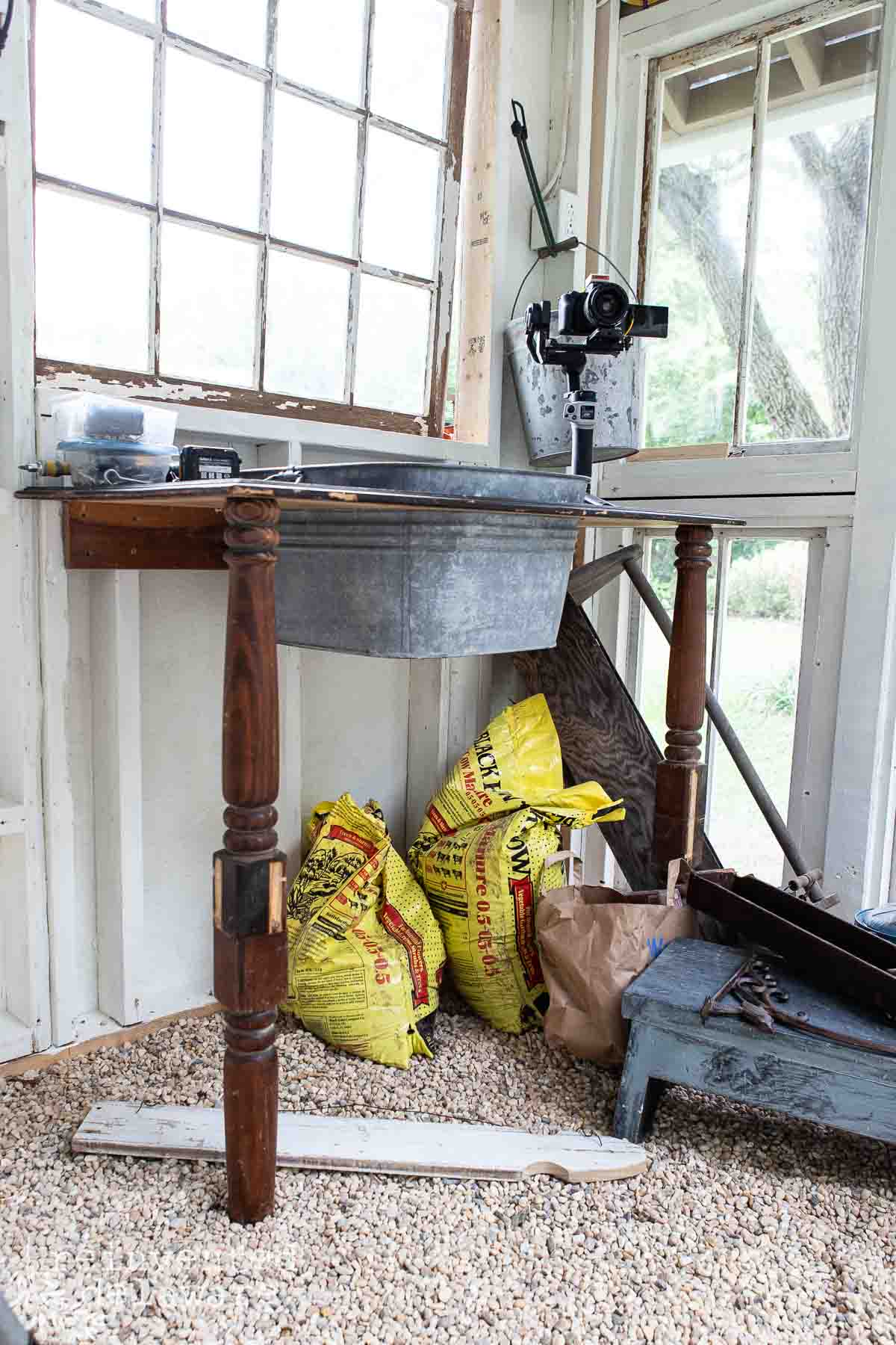 A gardening station in a sunlit garden shed featuring a vintage table with a metal top and wooden legs. On and under the table are gardening supplies, including several yellow bags of soil, a metal container, and a small blue wooden stool. Large windows provide ample natural light in a diy greenhouse.