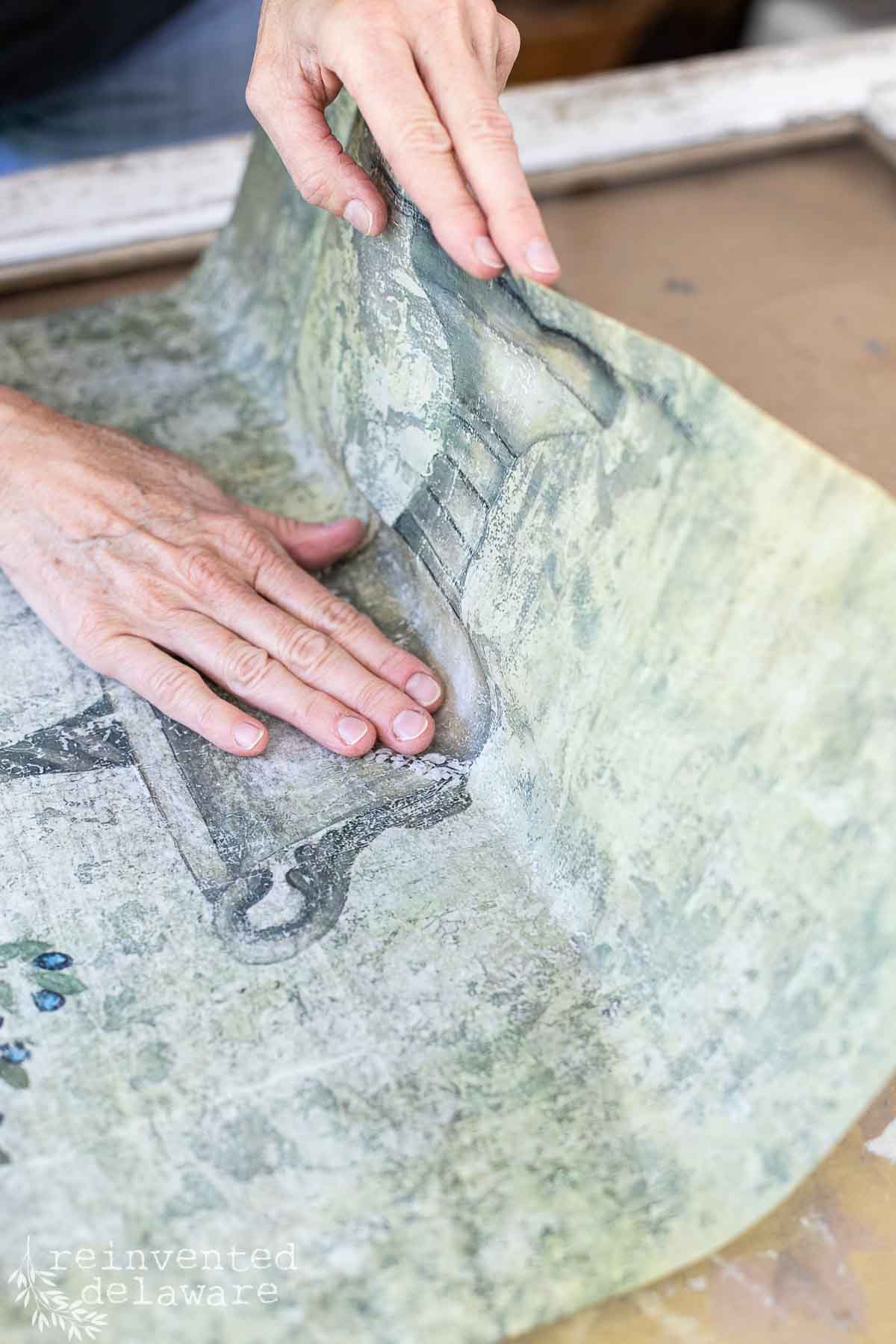 Close-up of a person pressing down a piece of vintage, worn-looking transfer paper onto a flat surface of a vintage window using their hands. The paper features an illustration of a stone planter. "Reinvented Delaware" is written in the lower left corner.