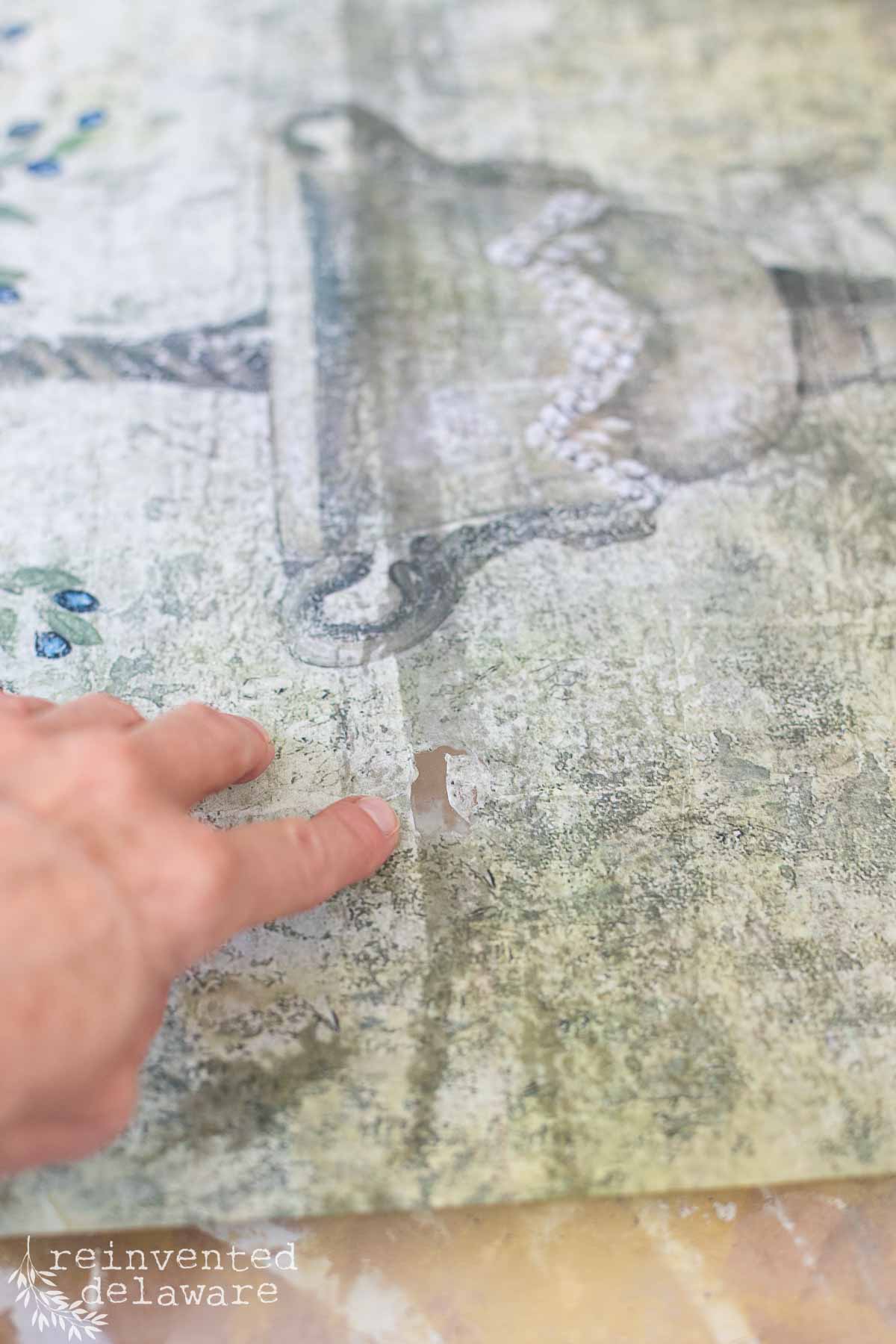 A close-up of a hand pointing at a small torn section on a vintage, distressed transfer. The poster has a faded design with some visible artistry, and there is faint text branding "reinvented delaware" in the bottom left corner of the image.