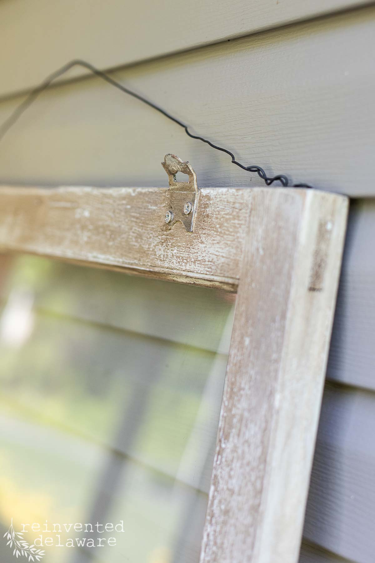 Close-up of an old, distressed wooden window frame hanging on a light gray, horizontally-slatted exterior wall. The frame is worn and chipped, with a metal hanger attached and a twisted wire for hanging. Text at the bottom reads "reinvented delaware.