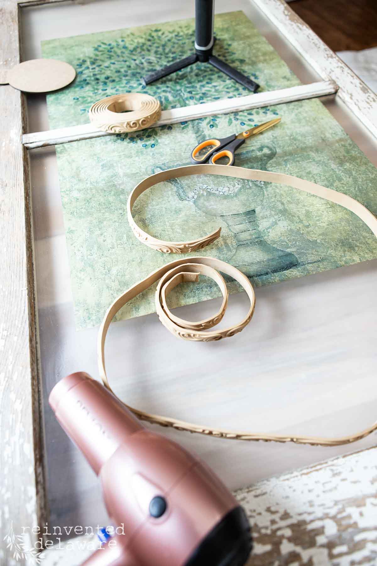An upcycle old window lies on a table with a pair of scissors, a hairdryer, and decorative trim. The picture has a green and blue scene with a potted plant. The trim is partially unrolled and decorated with intricate patterns.