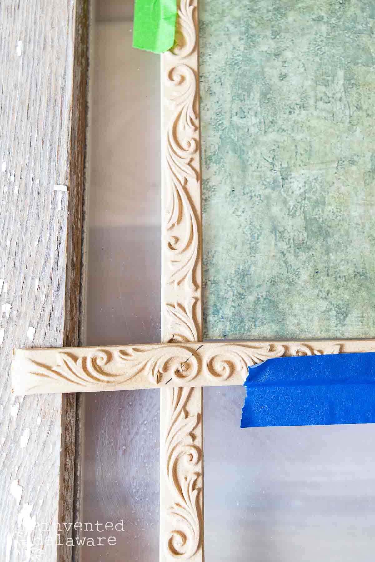 A close-up of a window frame with a section of decorative trim in progress. The trim has intricate, swirling designs and is partially taped with green and blue painter's tape for precision during installation or painting. The background shows a weathered wood grain texture.