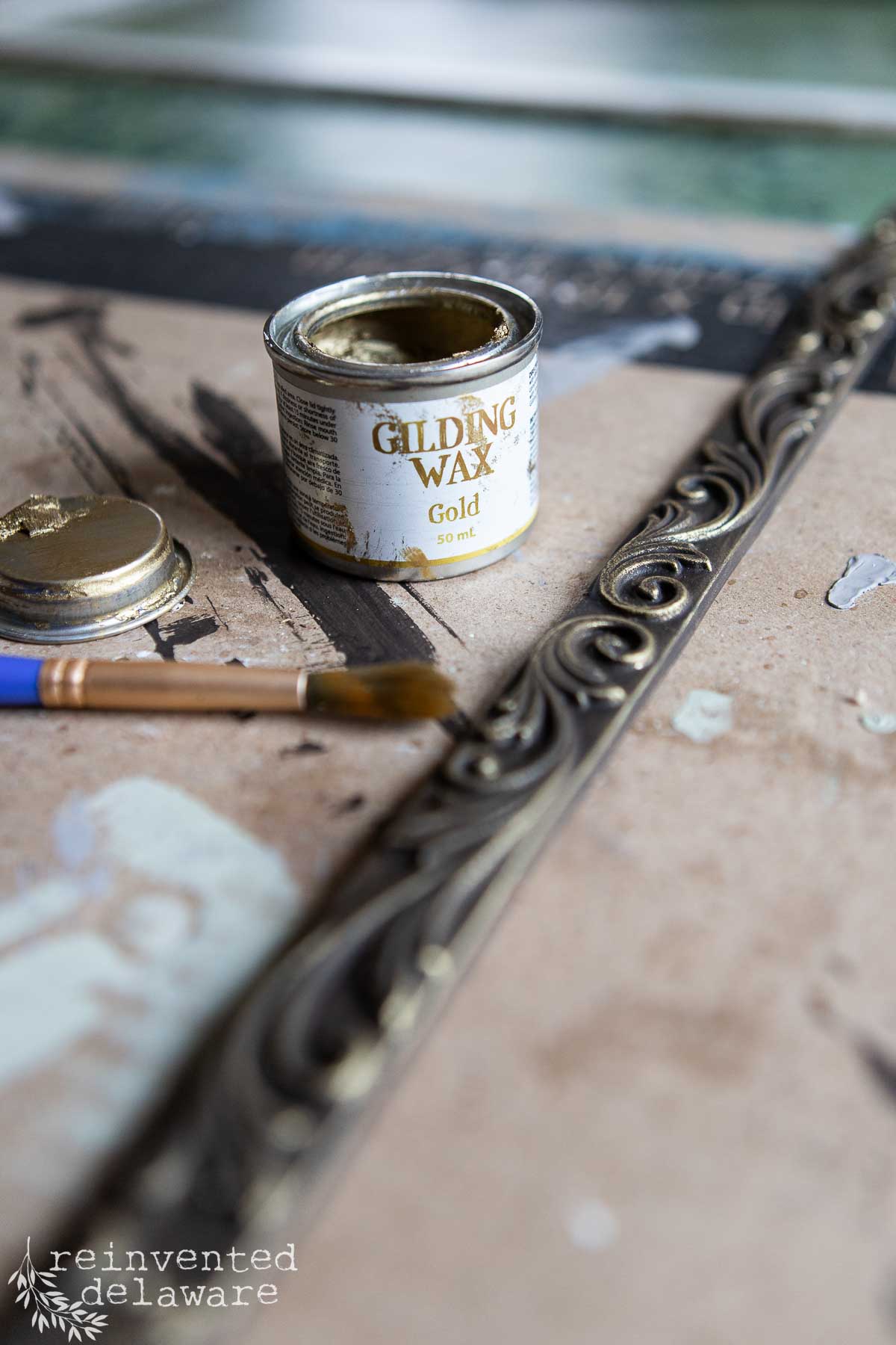 A small container of gold gilding wax, with its lid off, sits on a surface alongside a paintbrush and an ornate metallic strip. The surface is covered in scattered paint marks. The image is by Reinvented Delaware.