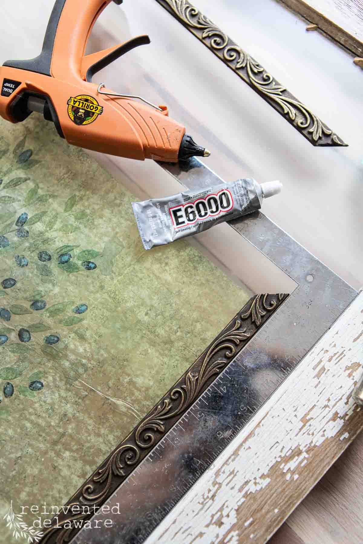 A hot glue gun, a tube of E6000 adhesive, and a piece of decorative trim are placed on an old window decorated with a green floral design. The edge of the glass is set in a distressed white wooden frame of an antique window, adorned with ornate trim corner pieces. The text "reinventeddelaware" is visible in the bottom left.