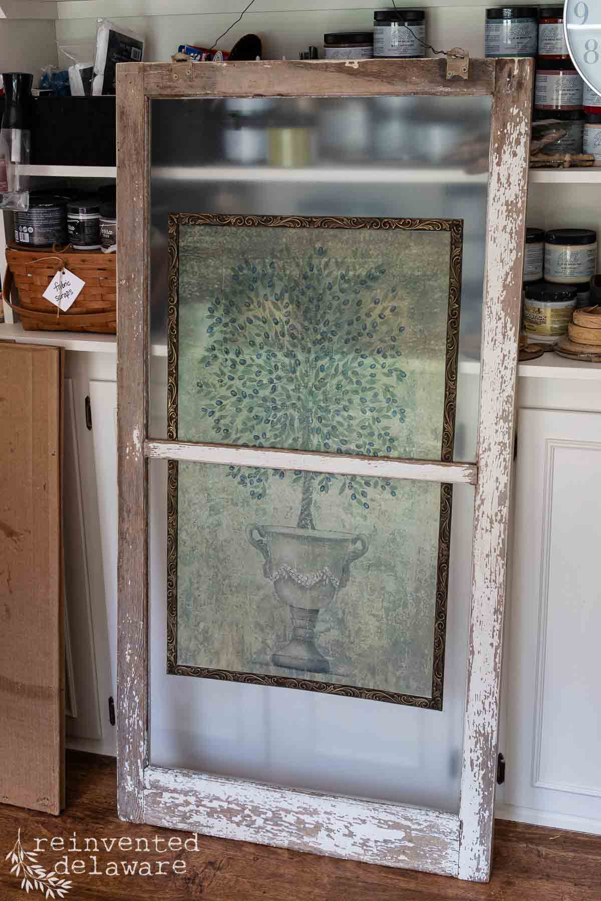 A rustic, old wooden frame with peeling white paint stands in a room. Inside the frame is a painting of a potted tree with lush green leaves. Shelves with various jars, cans, and art supplies are visible in the background.

.