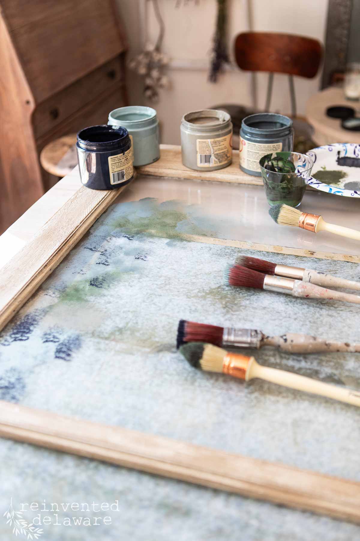 A table with several jars of paint, paintbrushes, and a old wooden window frame. The brushes and frame are resting on a glass surface with paint splotches. A chair and some hanging decorations are visible in the background. .