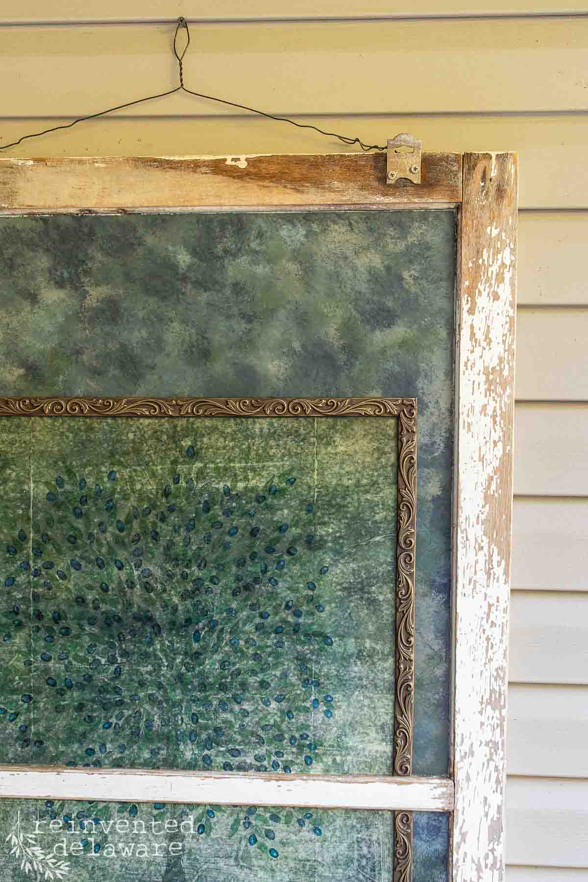 A rustic, weathered wooden window frame with peeling white paint hangs from a wire against a beige wall. Inside the frame is a textured, greenish-blue patterned artwork with small, scattered dots and an ornate border. The bottom corner features the text "reinventeddelaware.com.
