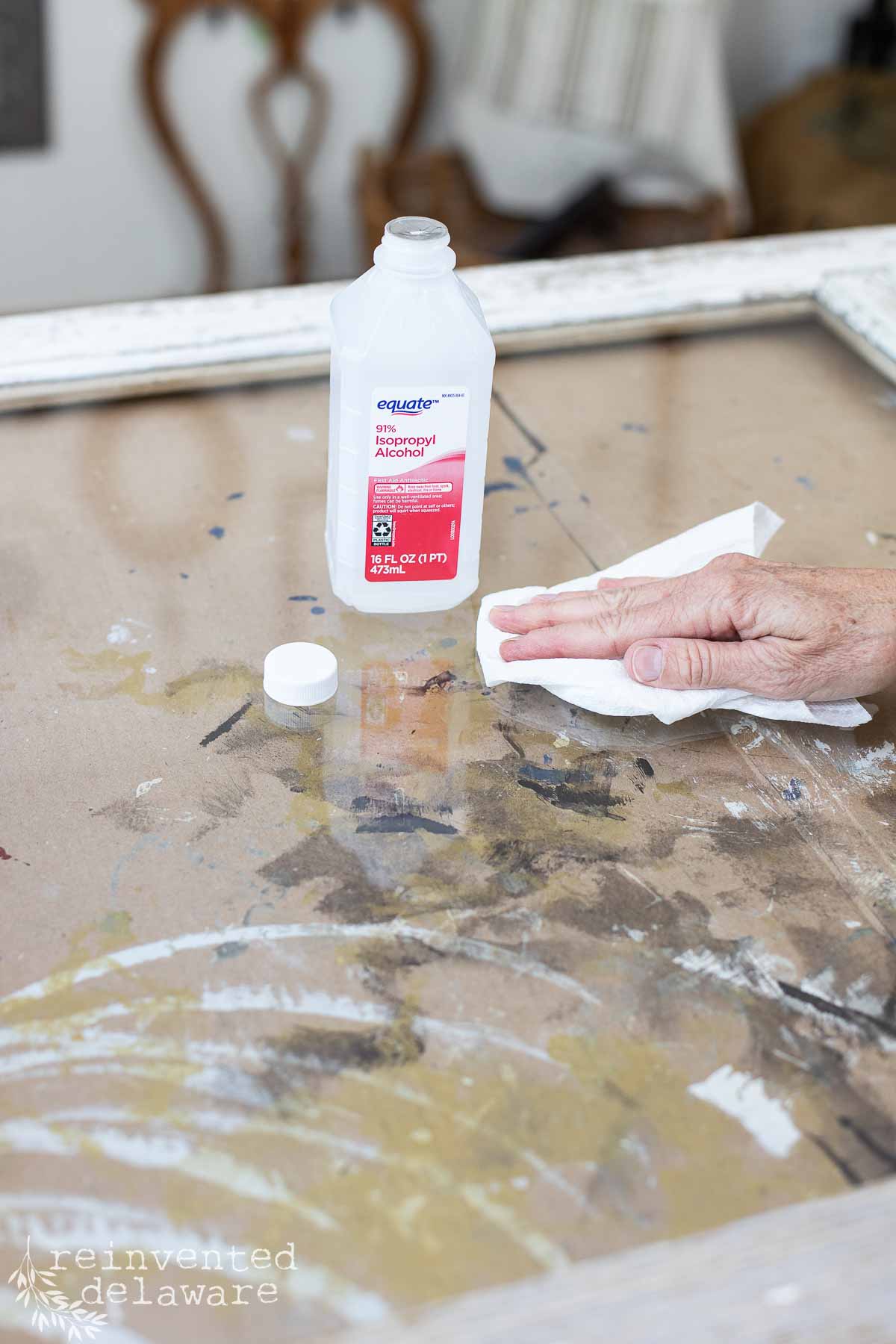 A person cleans a transparent surface with a white cloth. On the surface is an open bottle of Equate 70% Isopropyl Alcohol with the cap placed nearby. The surface has paint streaks and a wooden frame surrounds it. In the background is a blurry chair.