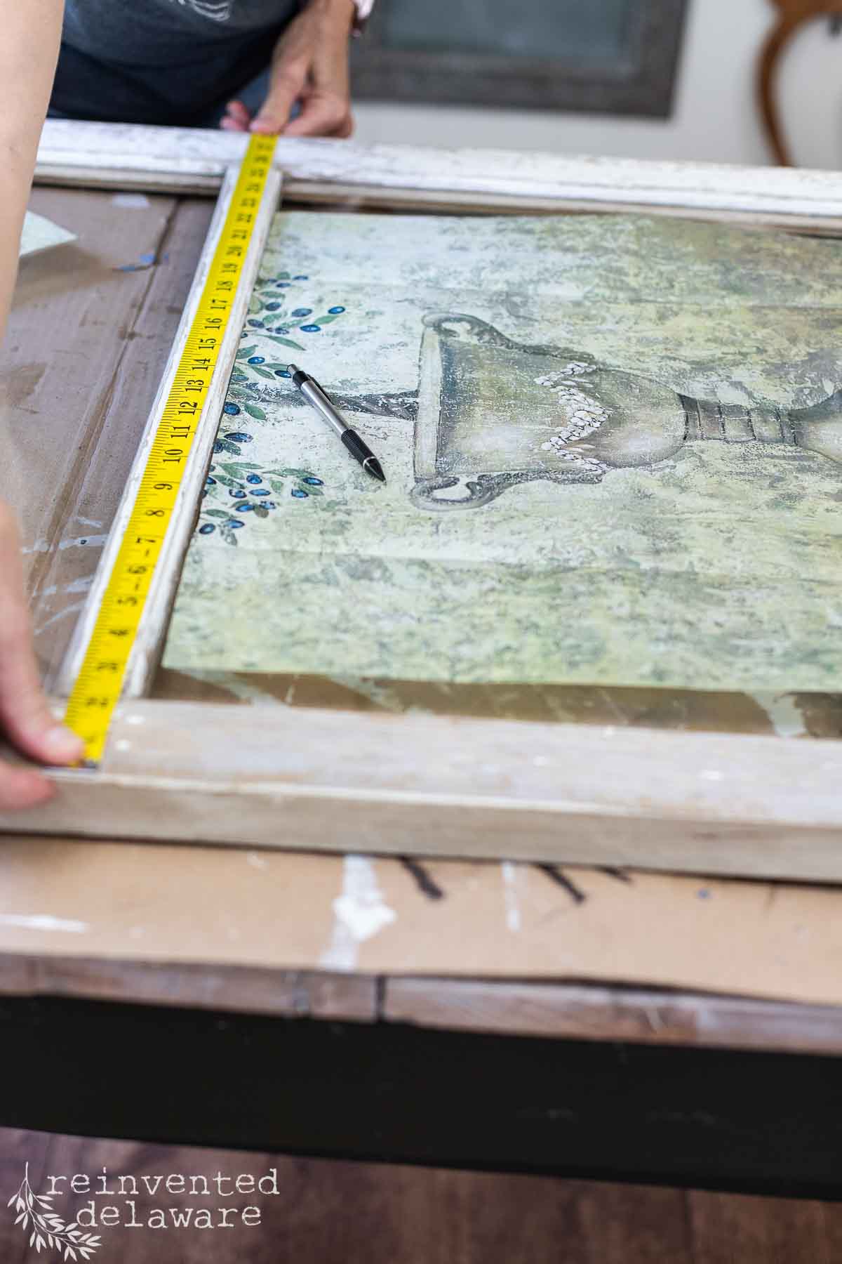 A person measures the edges of a large framed painting with a yellow measuring tape. The painting features a vase with floral designs. A marker rests on the artwork. The table below them has smudges of paint. The bottom left corner of the image shows the text "reinvented delaware.