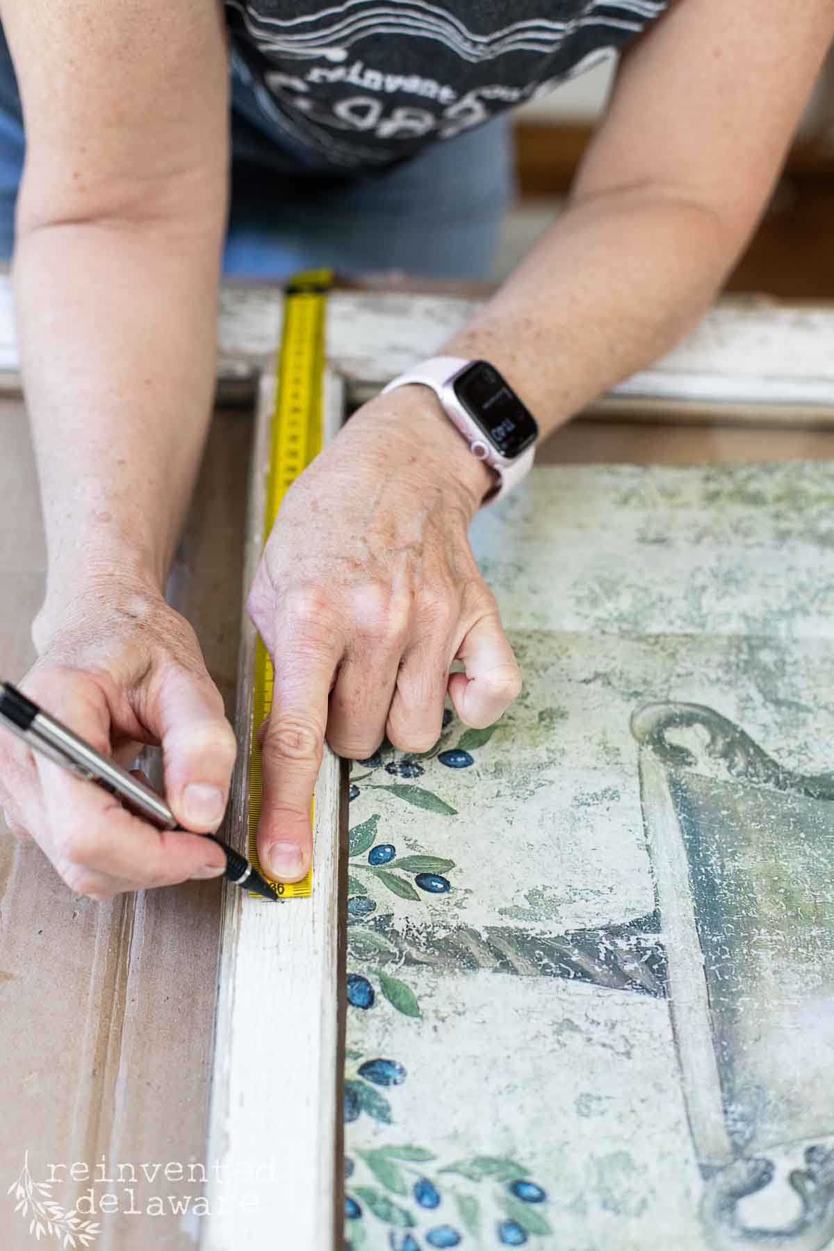 A person wearing a smartwatch is measuring the edge of a distressed wooden old window frame using a yellow tape measure and marking the measurement with a black pen. The frame has a decorative floral design. The person’s shirt is partially visible. Text in the bottom left corner reads "reinventeddelaware.com".