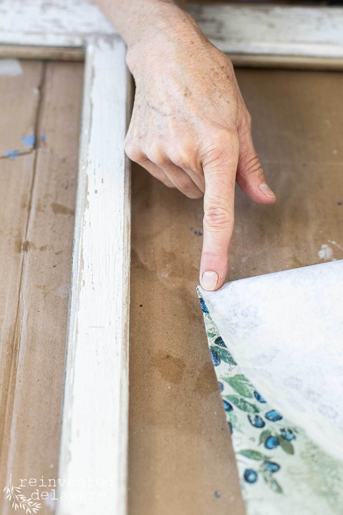 A person's hand is seen working on a vintage window wall decor DIY project. They are pointing at a corner of decorative paper or fabric in a floral pattern with blue and green colors, which is being applied to a wooden frame. The background is a brown cardboard surface.