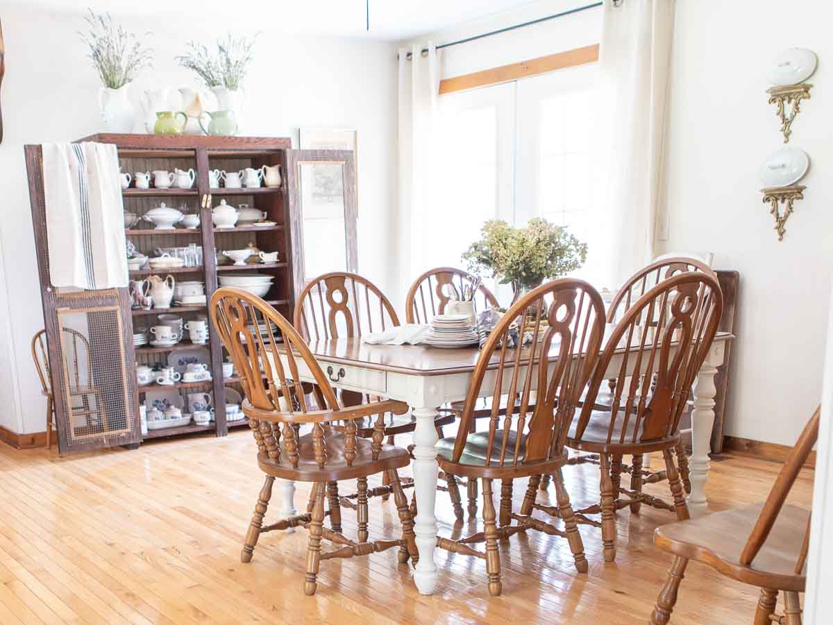 A bright dining room with wooden floors, a white dining table, and six wooden chairs surround it. A large cabinet displaying white dishware stands against the wall. Two wall-mounted shelves with antique plates and floral decorations are on the side wall.
