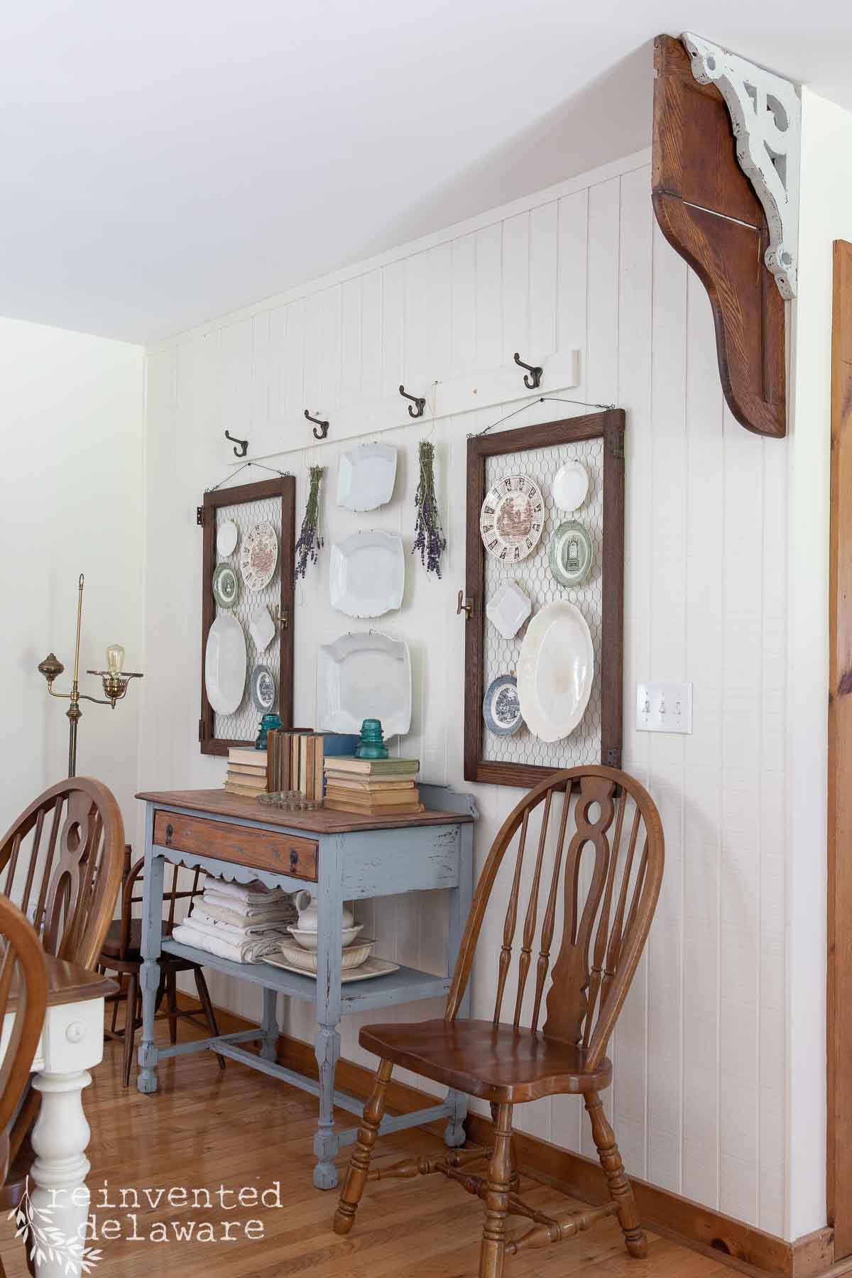 A cozy, vintage-styled dining room features two wooden chairs, a small wooden table with books and decor, and a wall adorned with frames filled with vintage ironstone plates. The room has wooden floors and white-paneled walls, with a decorative shelf bracket in one corner.