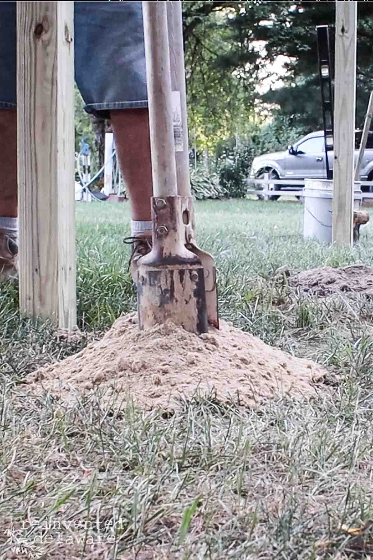 Close-up of a manual post hole digger standing vertically in a mound of dirt. The image also shows a person's legs wearing denim shorts and work boots. Wooden posts and a white car in the background can be seen in an outdoor grassy area.