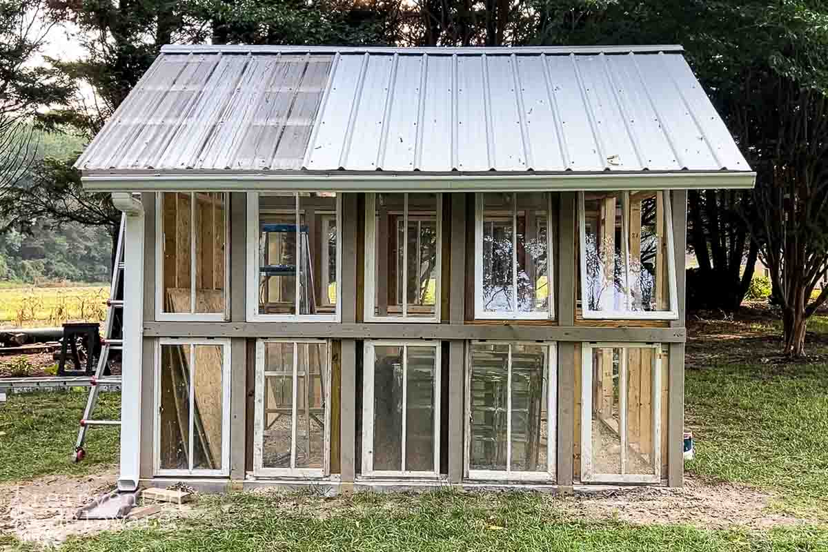 A partially constructed greenhouse stands on a grassy area with a white metal roof and multiple glass windows. A ladder leans on the left side inside, indicating ongoing work. Trees are visible in the background.