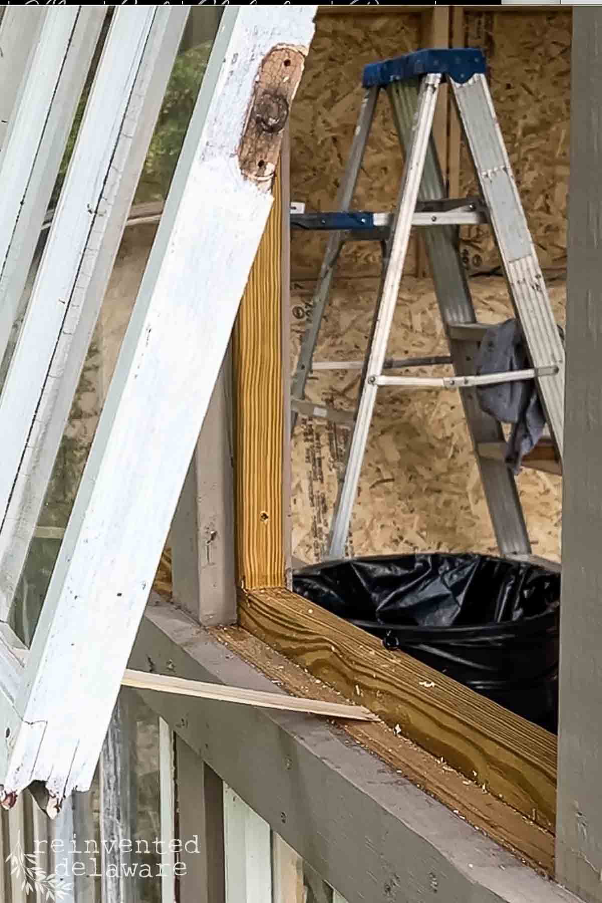 An opened window with peeling white paint beside a wooden frame under construction. A blue stepladder and a black trash bag are visible inside the structure. The background features partially installed plywood walls.