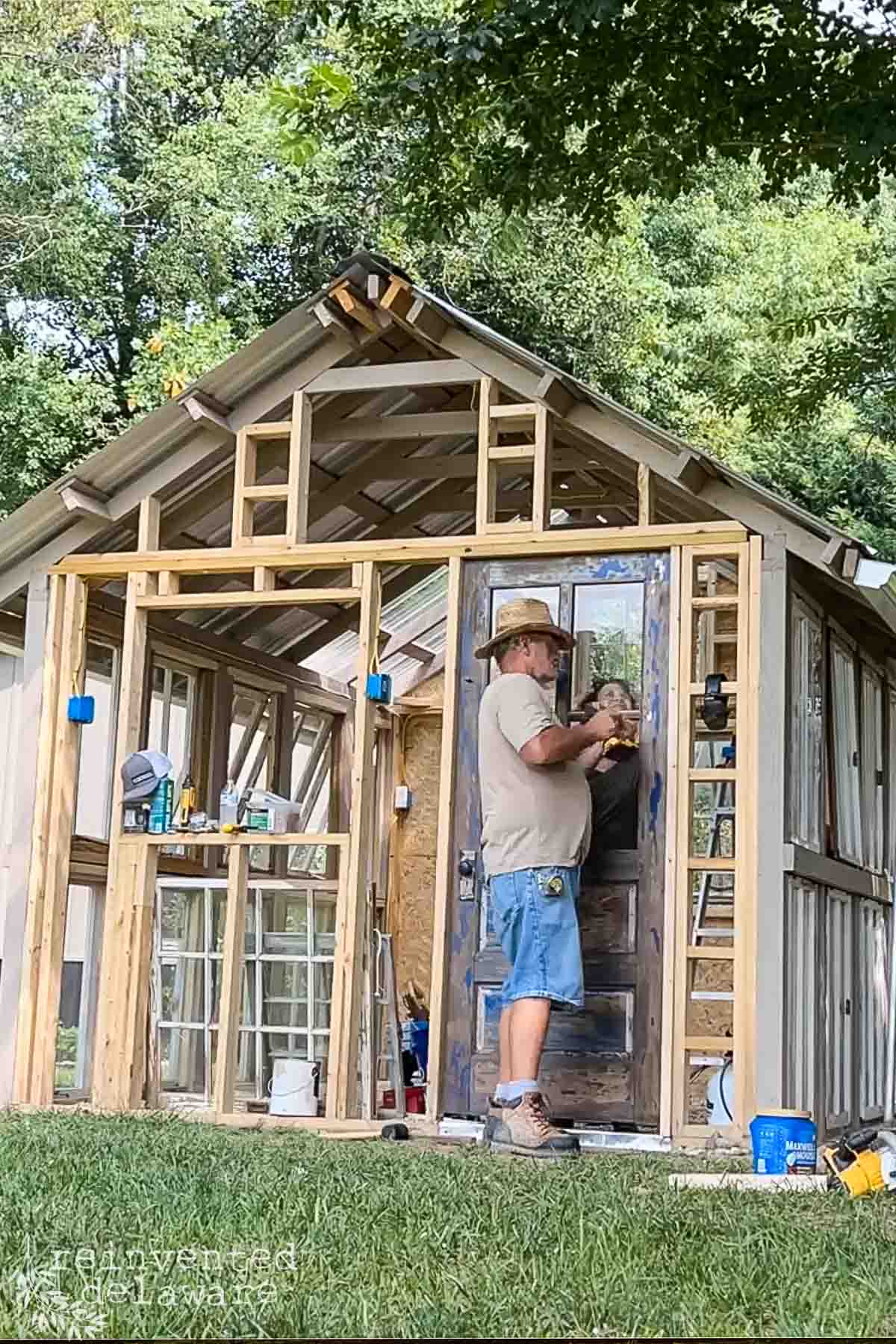 A man wearing a straw hat and denim shorts is working on the partially constructed wooden framework of a small building, possibly a greenhouse or shed. He is using a tool on a door. Various construction materials and tools are scattered around him. Trees and greenery are in the background.