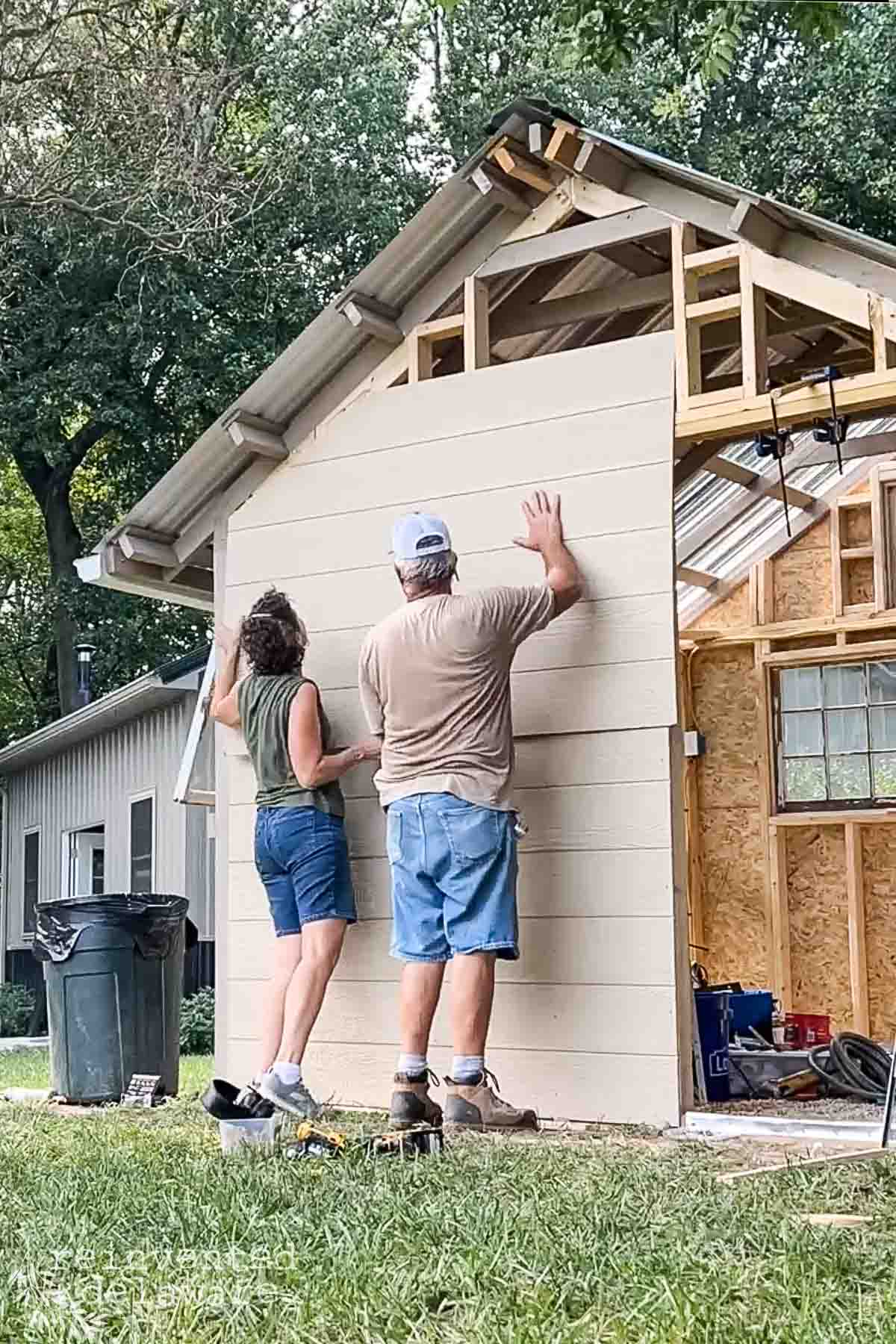 Two people are installing horizontal siding on a small house or shed. They are positioned on either side, holding up a large panel of siding. The structure has a partially finished exterior and an exposed framework at the eaves. Tools are visible on the ground.