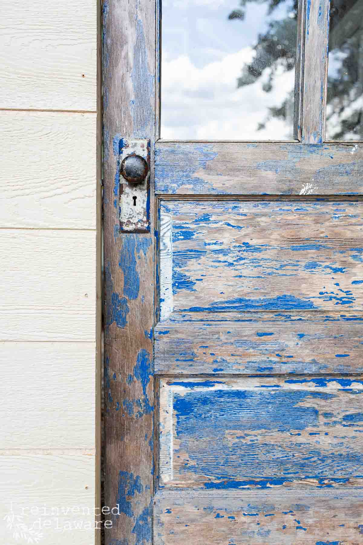 A close-up of a weathered wooden door with peeling blue paint. The door features a rustic, metal doorknob and glass panes at the top. The door is framed by light-colored siding. The texture and layers of the worn paint give it a vintage, distressed appearance.