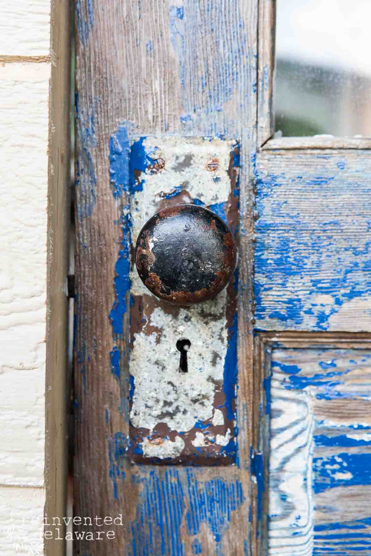 A close-up of an old, weathered door with peeling blue paint on the wooden surface. The door features a rusted, antique doorknob and keyhole mounted on a patina-covered metal plate, showcasing signs of age and wear. "reinvented delaware" is written in the bottom-left corner.