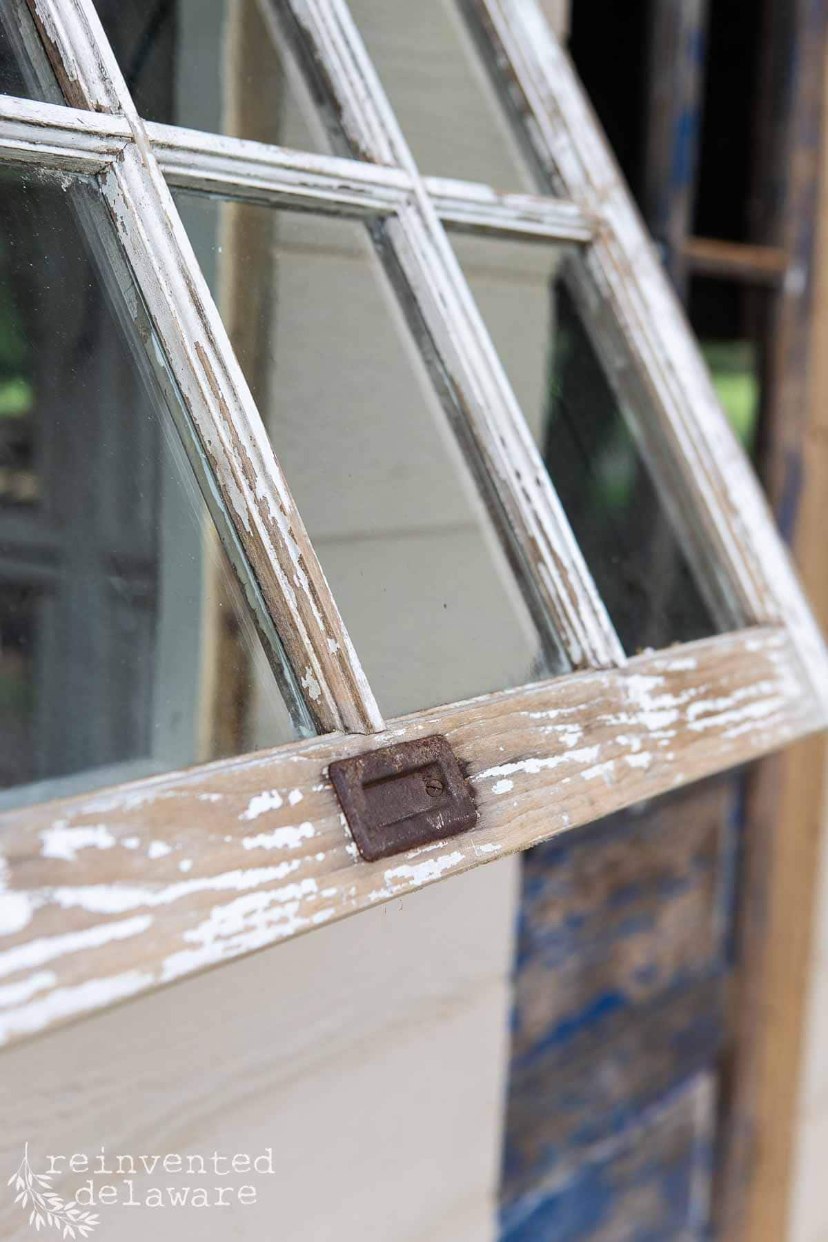 Close-up of a weathered, vintage window with a wooden frame and diamond-shaped panes. The paint is chipped and peeling, revealing a rustic and aged texture. A metal latch is visible on the window. The text "reinvented delaware" is in white in the bottom left corner.