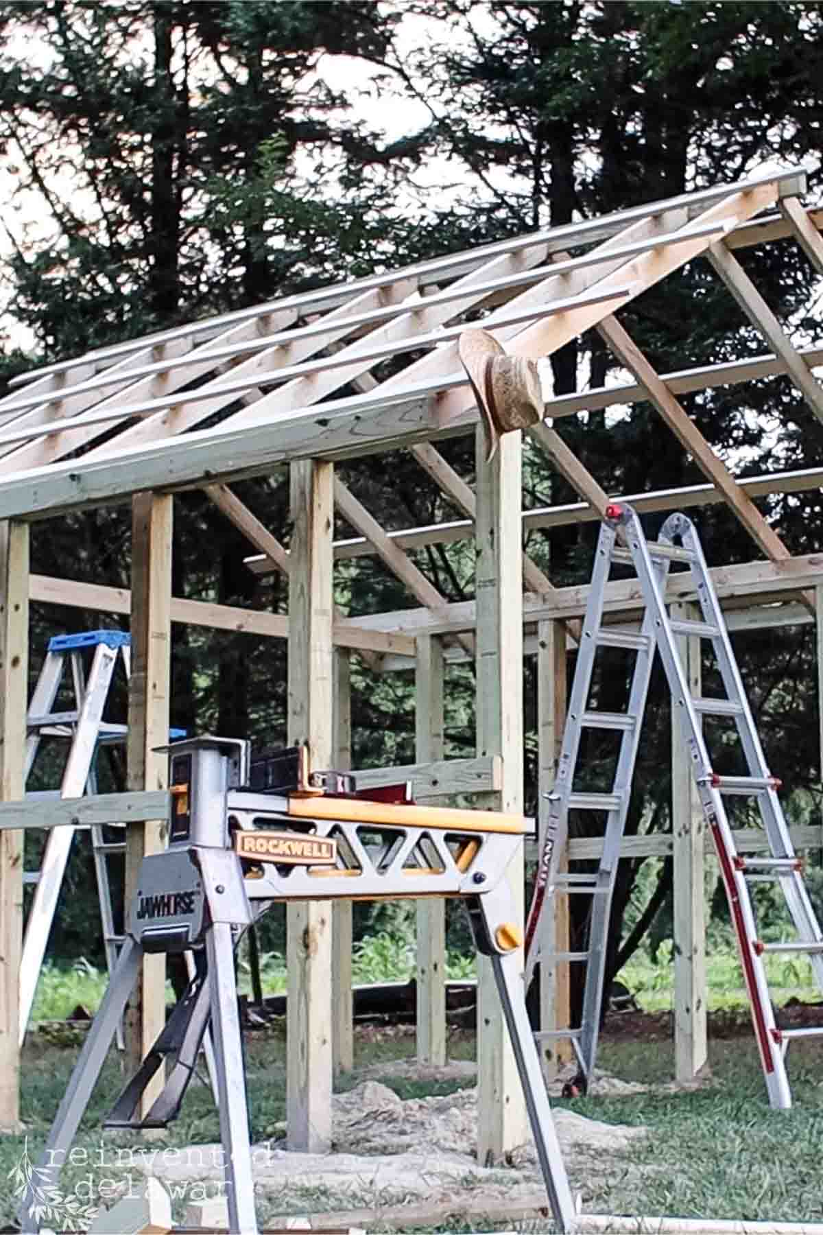 A partially constructed wooden shed frame stands amidst greenery and trees. Two ladders are propped up against the frame, and a workbench with a saw sits nearby. A straw hat rests on one of the beams.