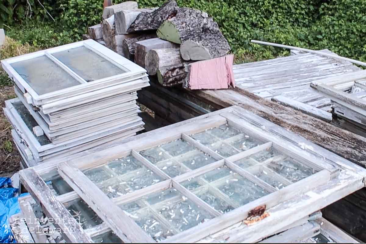 A pile of old, weathered wooden frames and window panes is stacked outdoors. The frames are peeling with white paint and rest amongst pieces of rough-cut wood and logs. Lush green vegetation can be seen in the background.