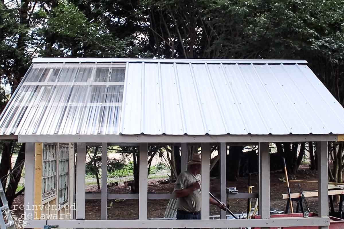 A person works on constructing a small shed with a partially transparent roof in a wooded area. The shed frame is white, and the roof is composed of corrugated metal and clear panels. Trees and various tools are visible in the background.