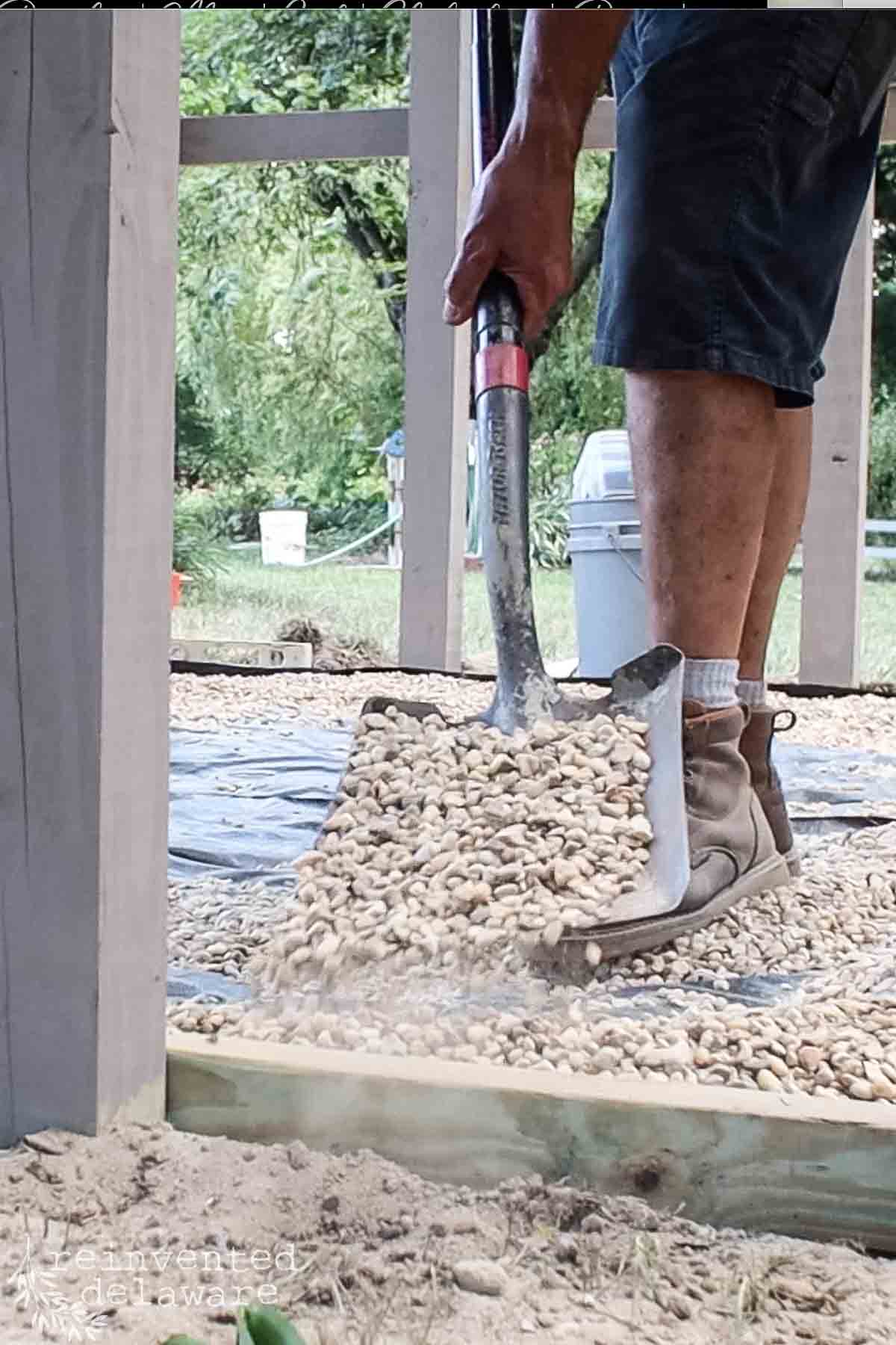 A person wearing boots and shorts is spreading gravel with a shovel on a leveled surface. The scene is set outdoors, with greenery visible in the background and construction materials around the area.
