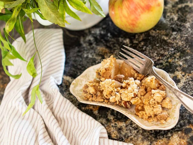 A plate of apple crumble with a fork and spoon rests on a marbled countertop, accompanied by a scoop of blueberry frozen yogurt. A striped cloth is nearby, with green leaves and a whole apple in the background, creating a cozy kitchen atmosphere.
