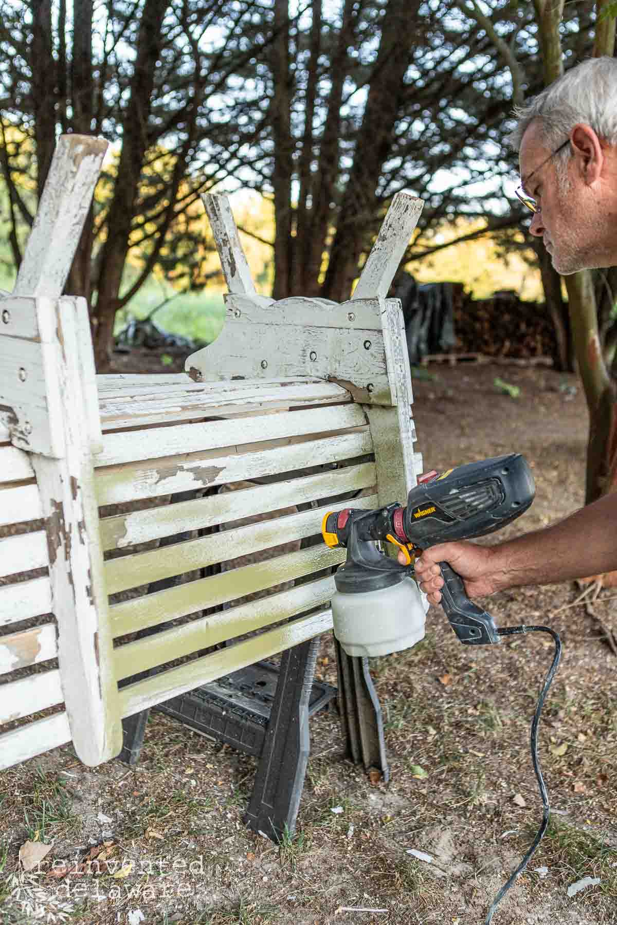 A man wearing glasses uses a Wagner spray paint tool to paint an inverted wooden bench in an outdoor area surrounded by trees. The bench appears weathered, and some sections are chipped and being covered with fresh paint.