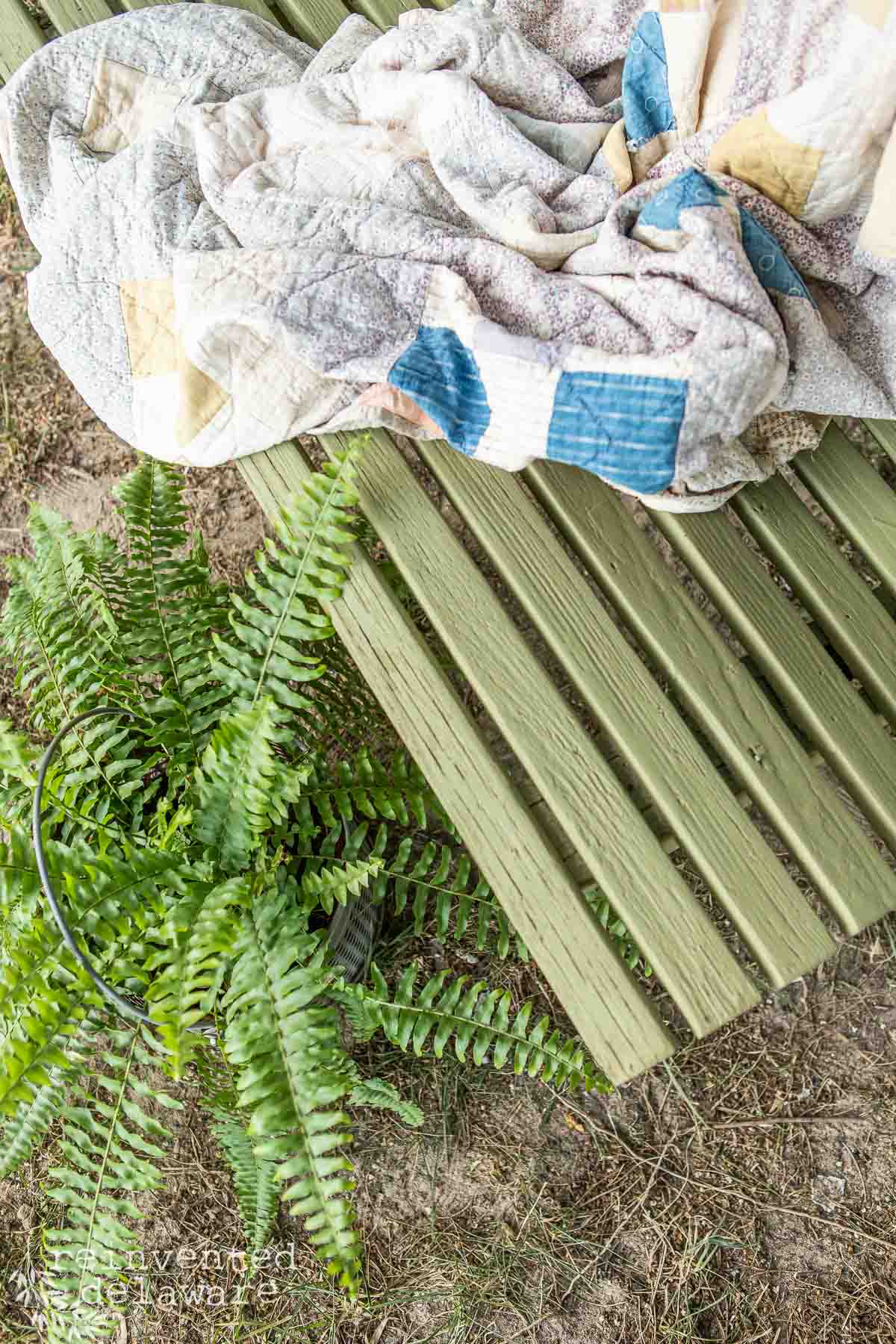 Painted outdoor wooden furniture showing the detail of the paint on a wood bench. Also displayed with an antique quilt and a potted fern plant.